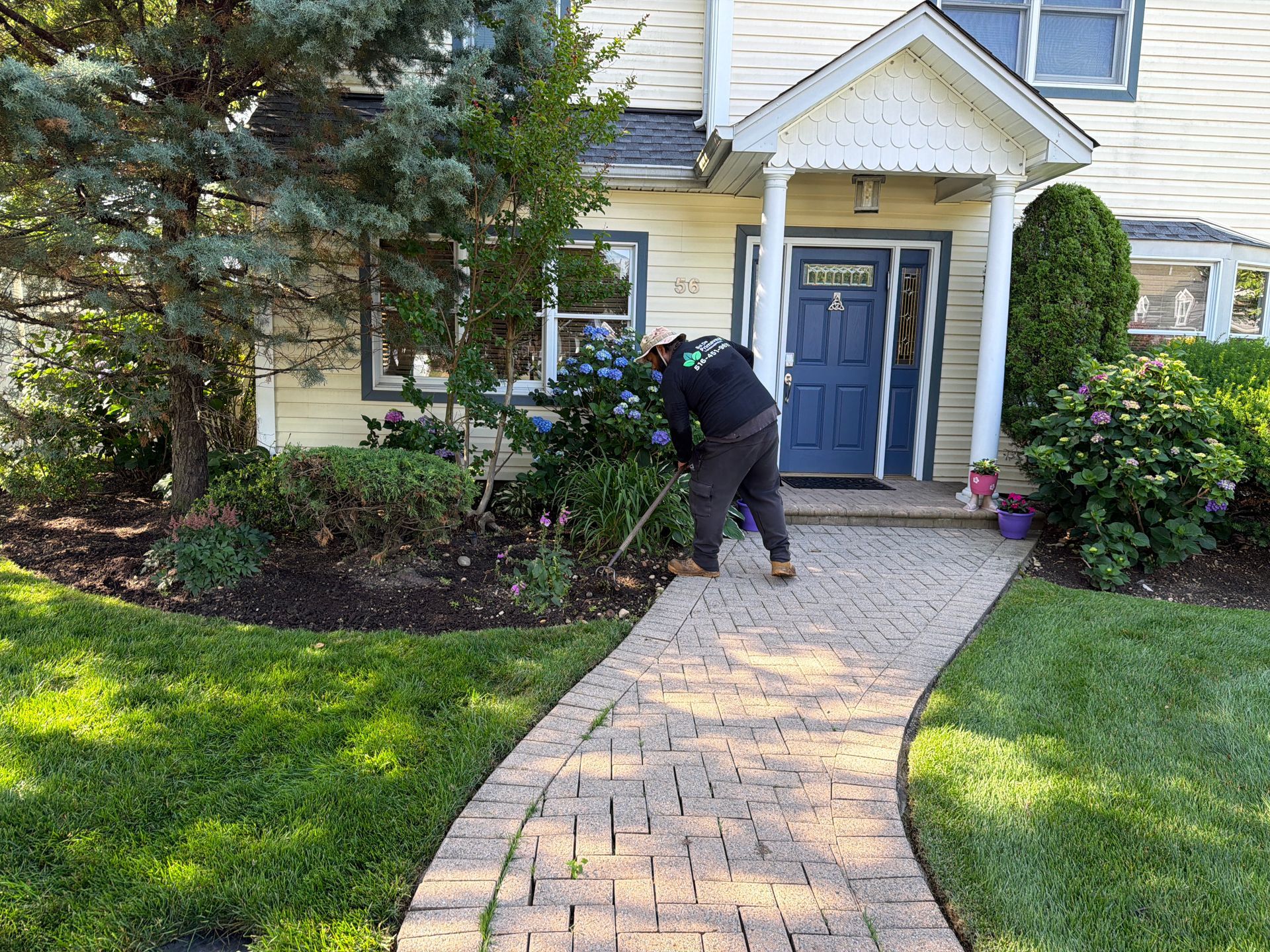 Person trimming shrubs beside a yellow house with a blue door and brick walkway