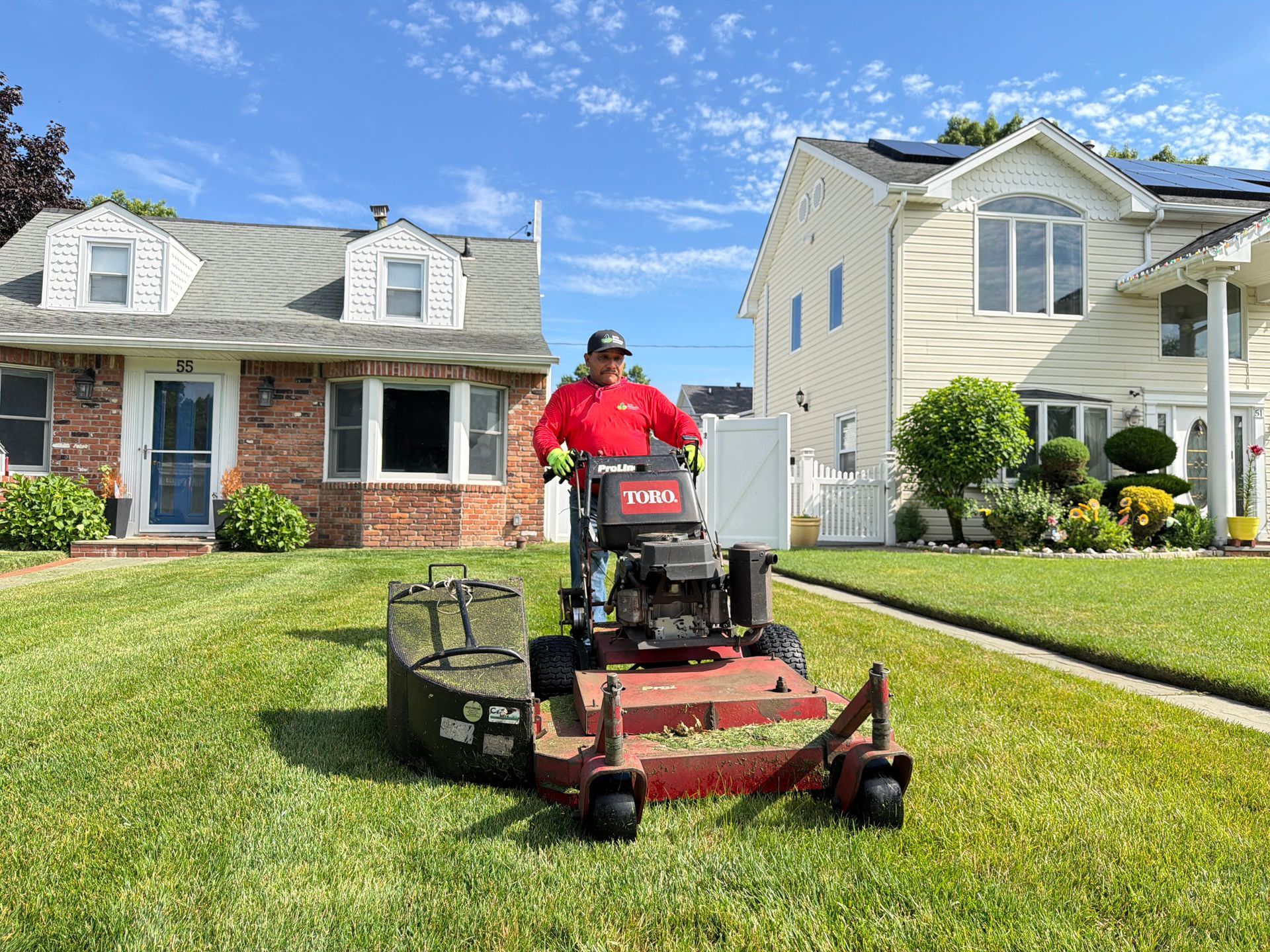 Man mowing a lawn with a red riding mower in front of suburban houses under a blue sky