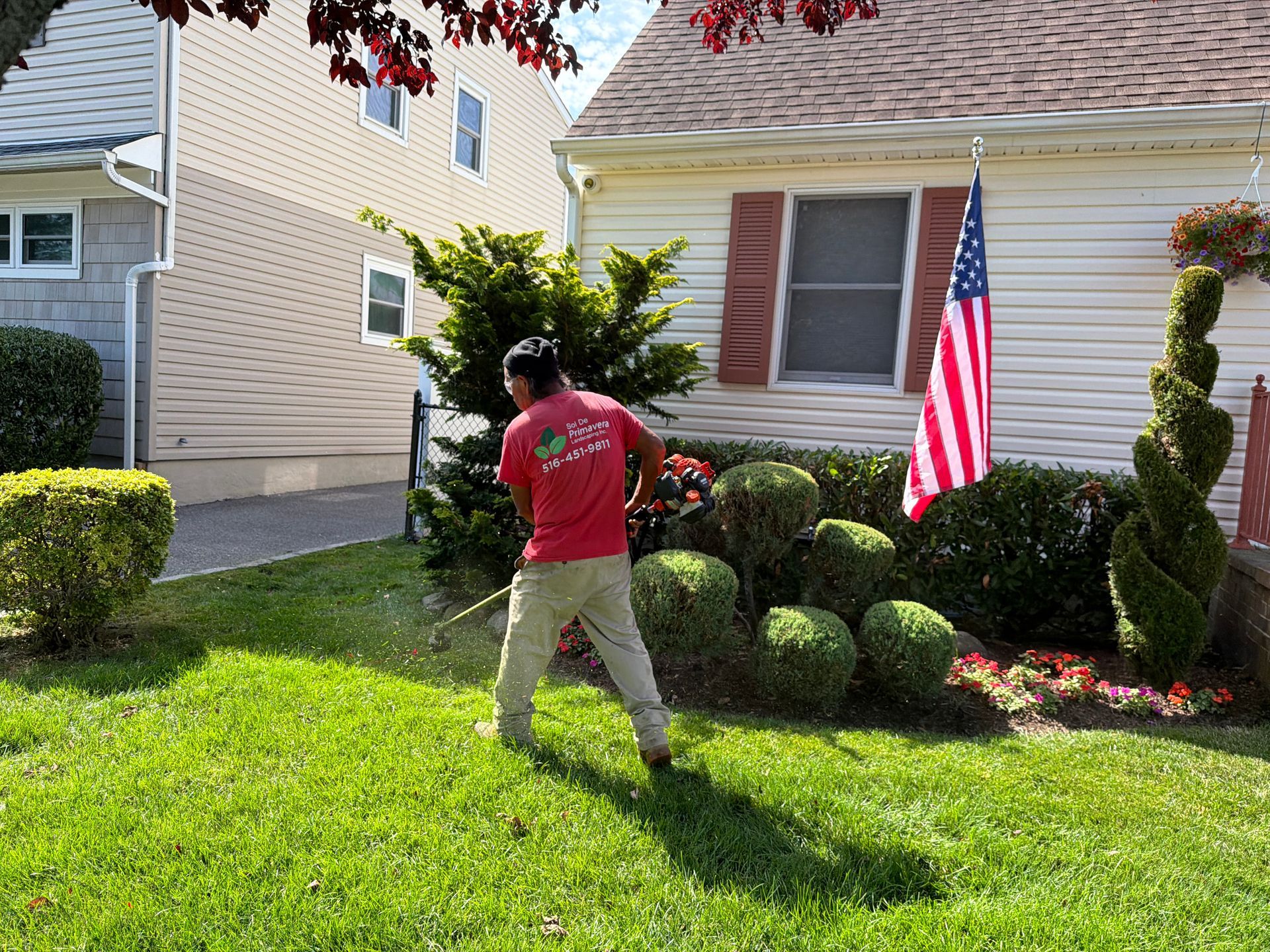 Person mowing a front lawn beside a house with an American flag and trimmed shrubs.