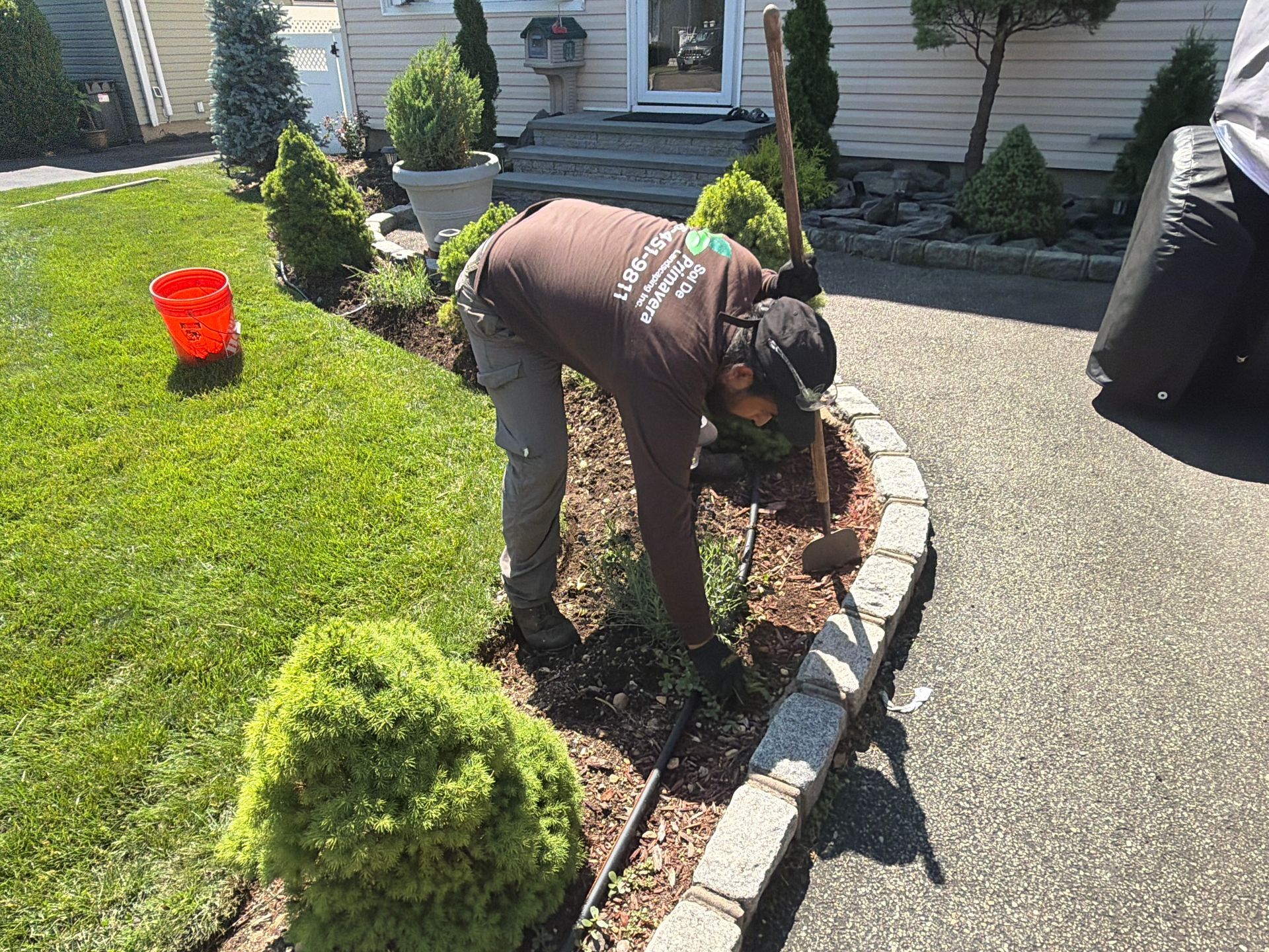 Gardener trimming shrubs along a curved stone border beside a driveway.