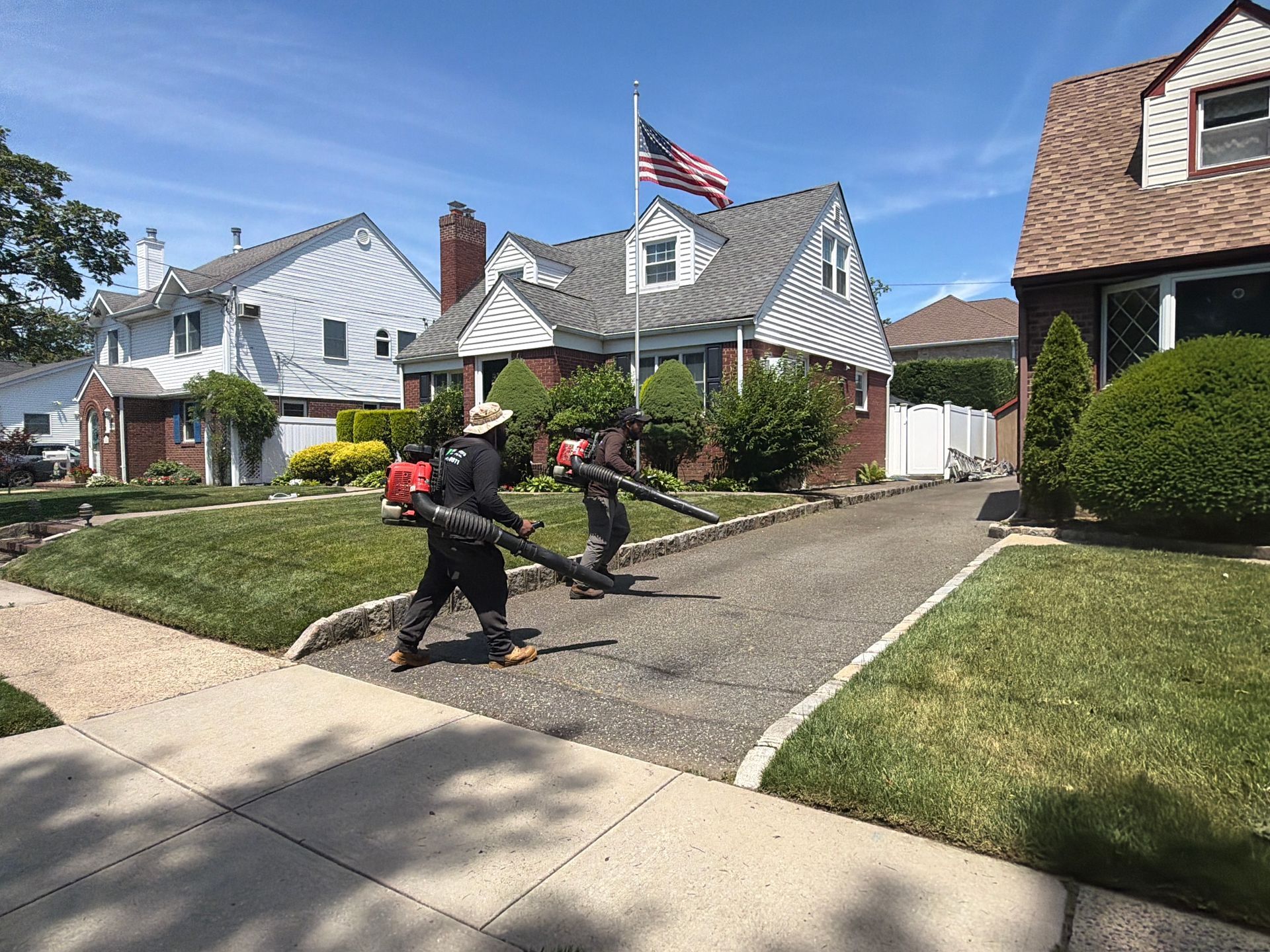 Two workers using leaf blowers on a suburban driveway beside houses with an American flag.