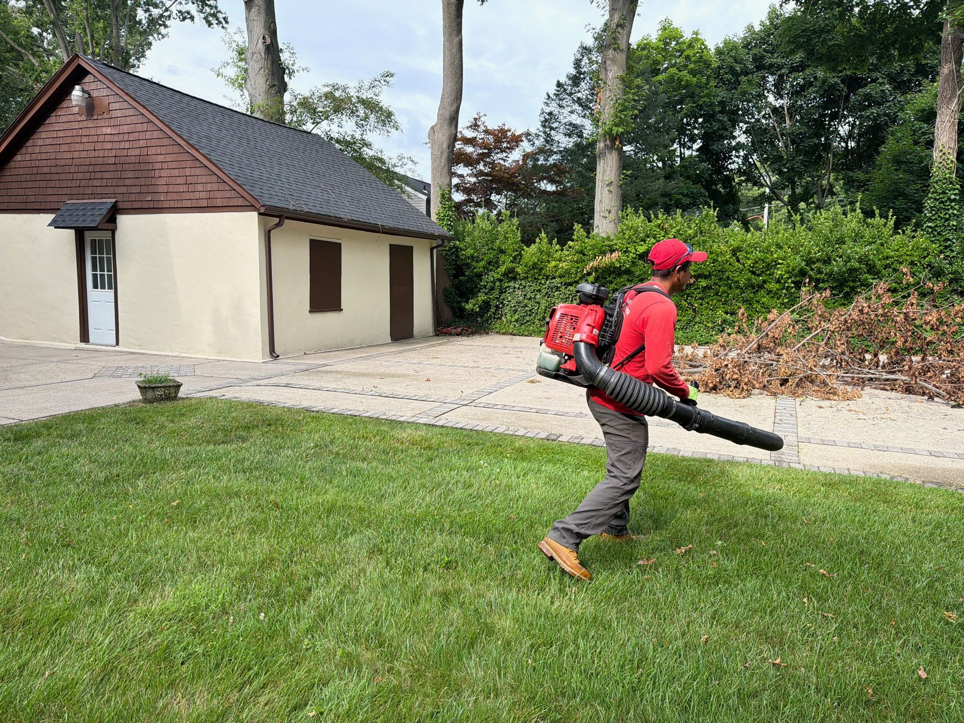 Person in red walks with a leaf blower across a grassy yard near a small building.