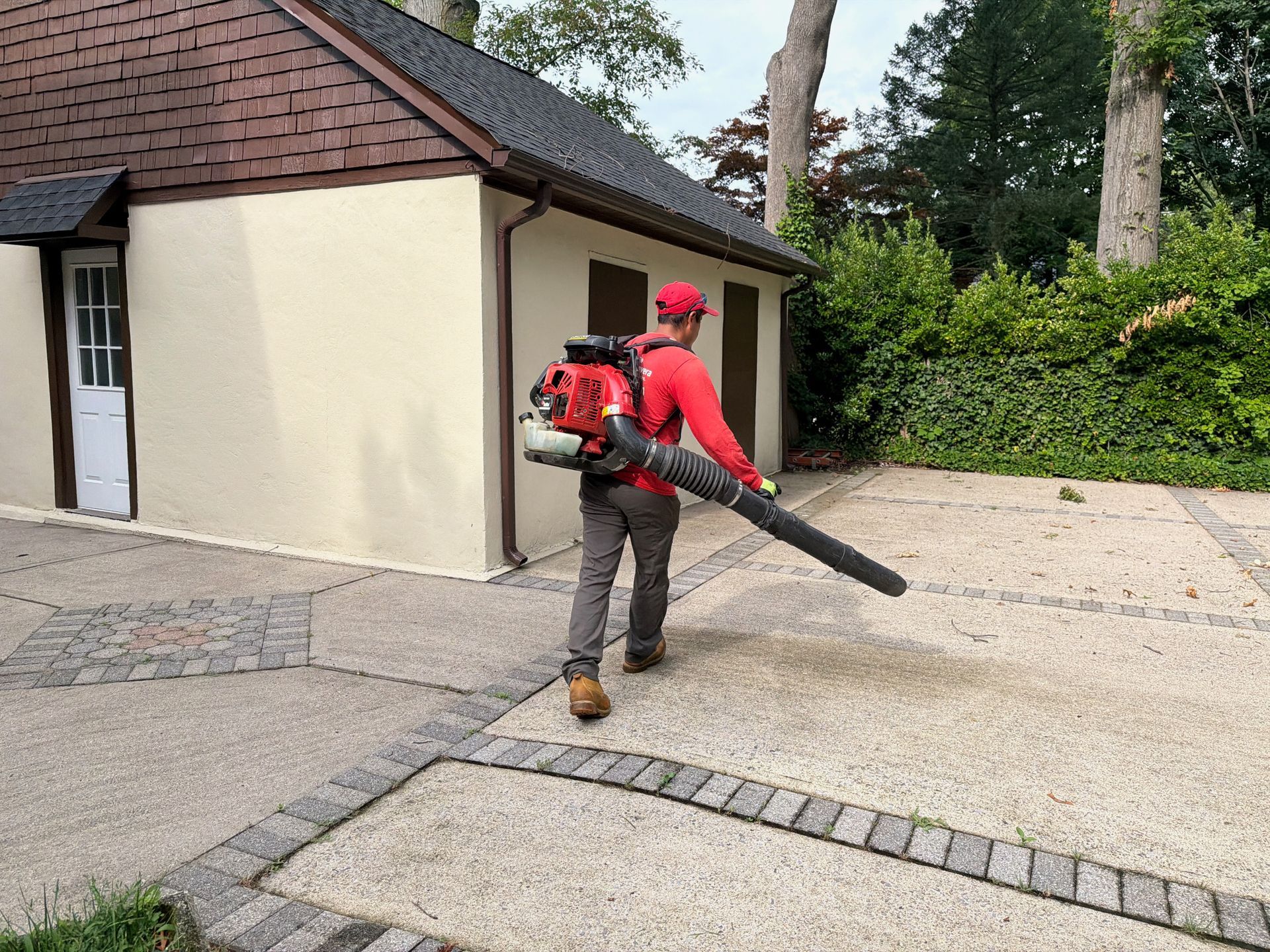 Worker using a red leaf blower near a house and driveway, clearing debris outdoors.