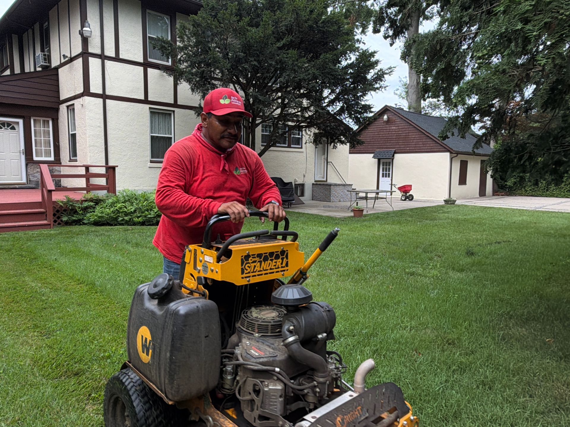 Person in red jacket and cap operating a yellow lawn machine on a grassy yard near houses.