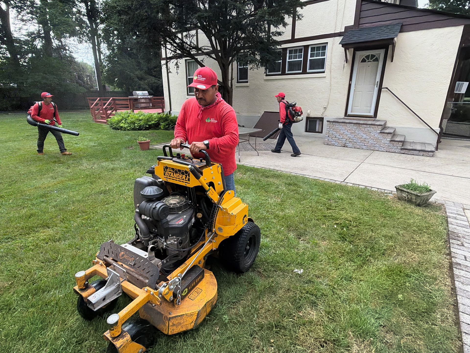 Worker mowing grass with a yellow ride-on mower near a house while others use leaf blowers.