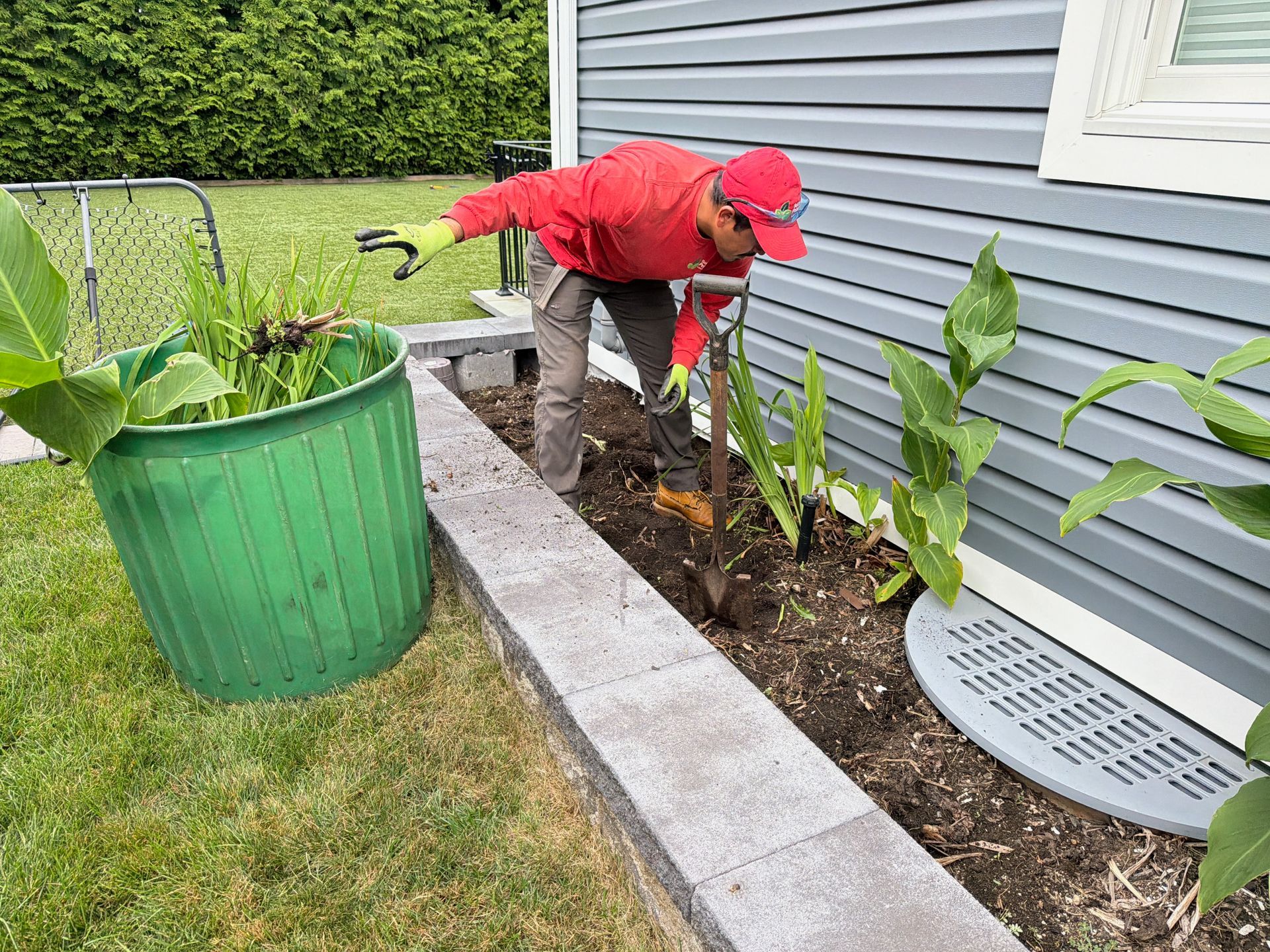 Person in red cap and shirt planting in a raised garden bed beside a house.