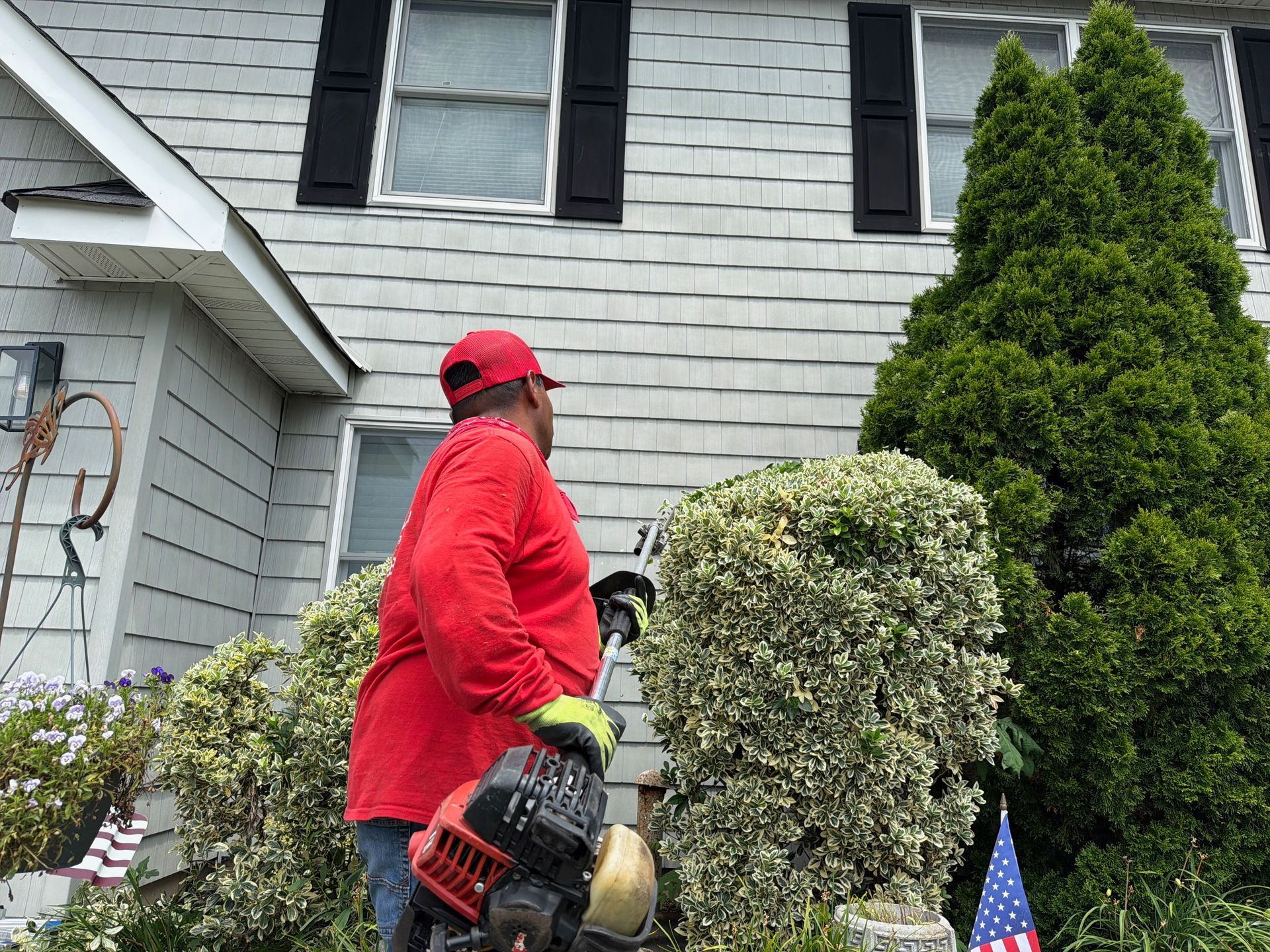 Person in red jacket and cap using a leaf blower outside a house with shrubs and an American flag.