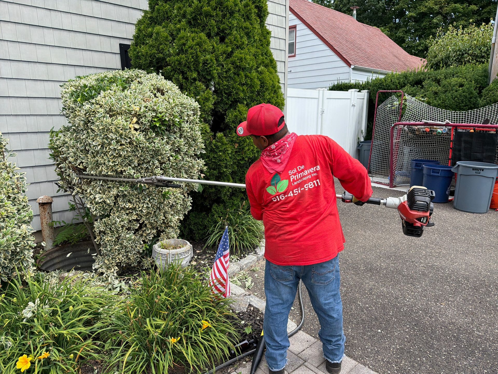 Person in red shirt using a string trimmer beside a house, with a small American flag in the yard.
