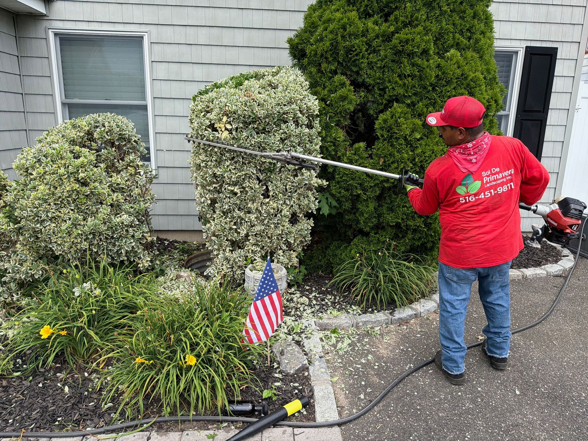 Worker in red shirt spraying shrubs beside a house, with an American flag in the garden.