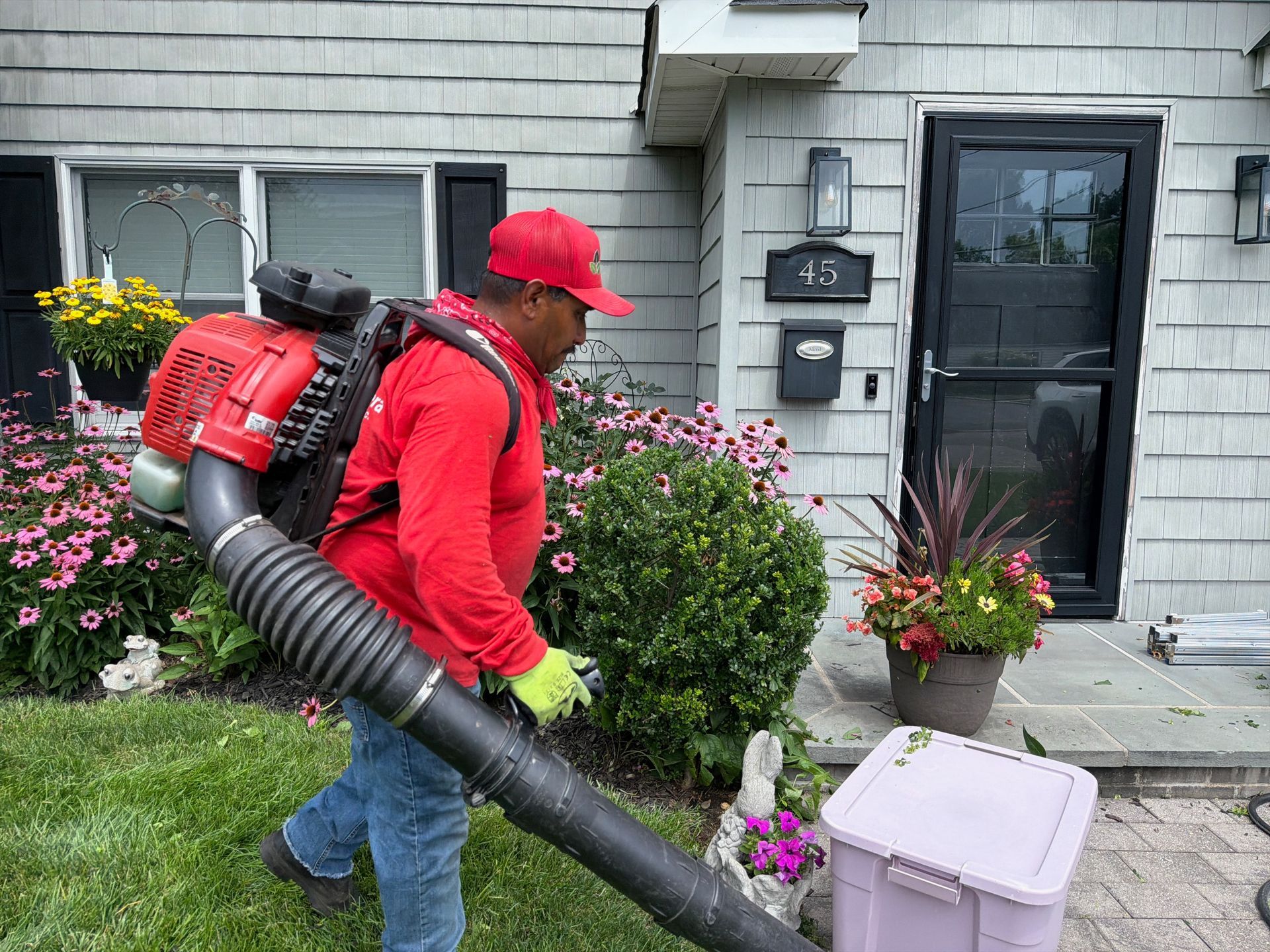 Worker in red using a backpack leaf blower beside a house walkway and front door