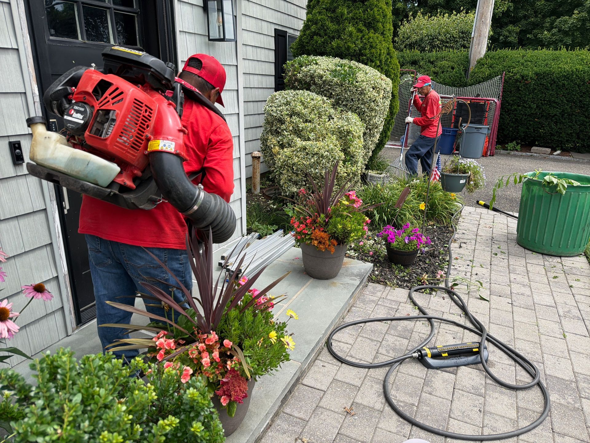 Landscapers using red leaf blowers along a house walkway beside potted flowers and trimmed hedges