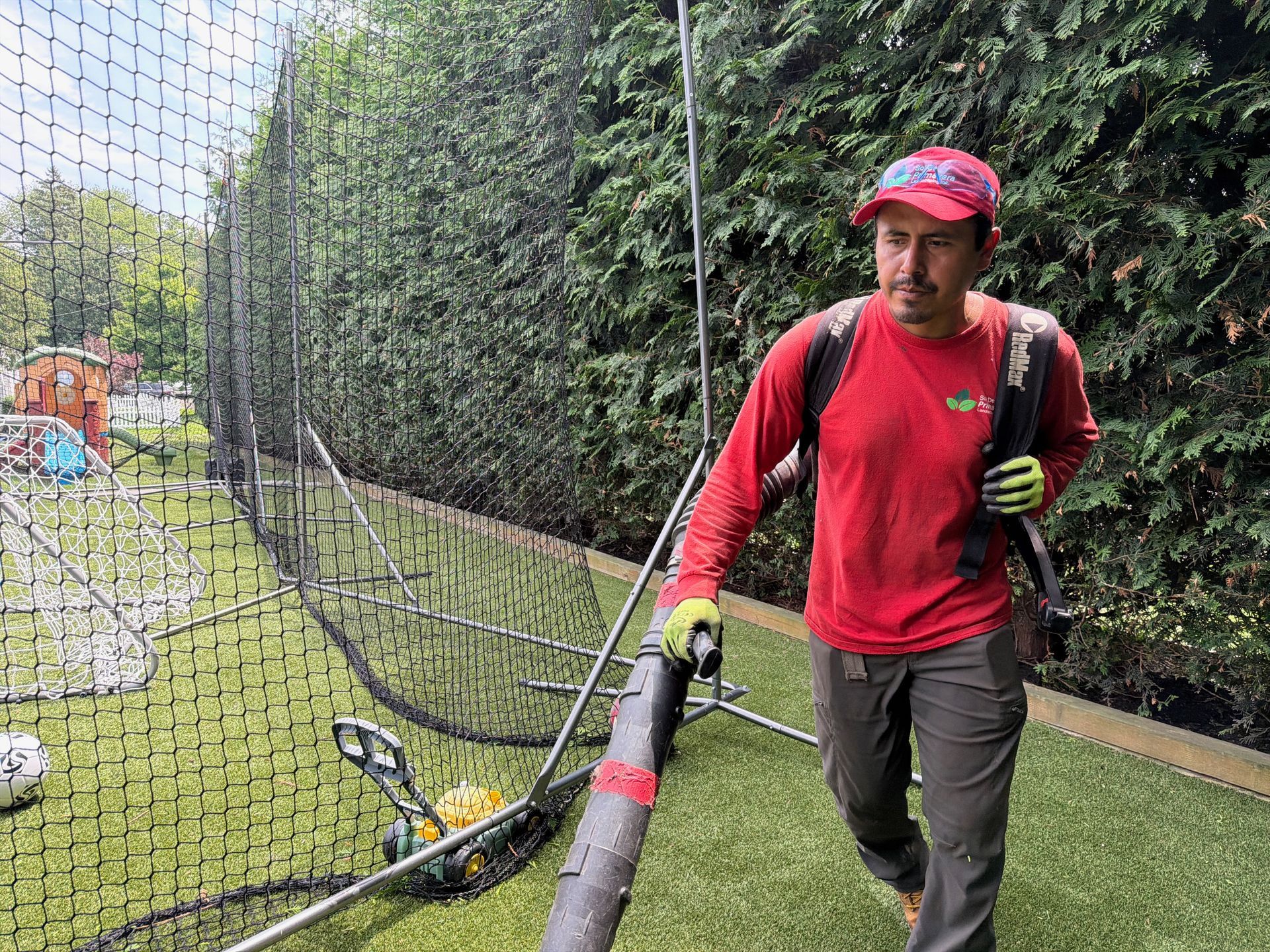 Worker in red shirt mowing a grassy sports field beside tall netting and hedges.