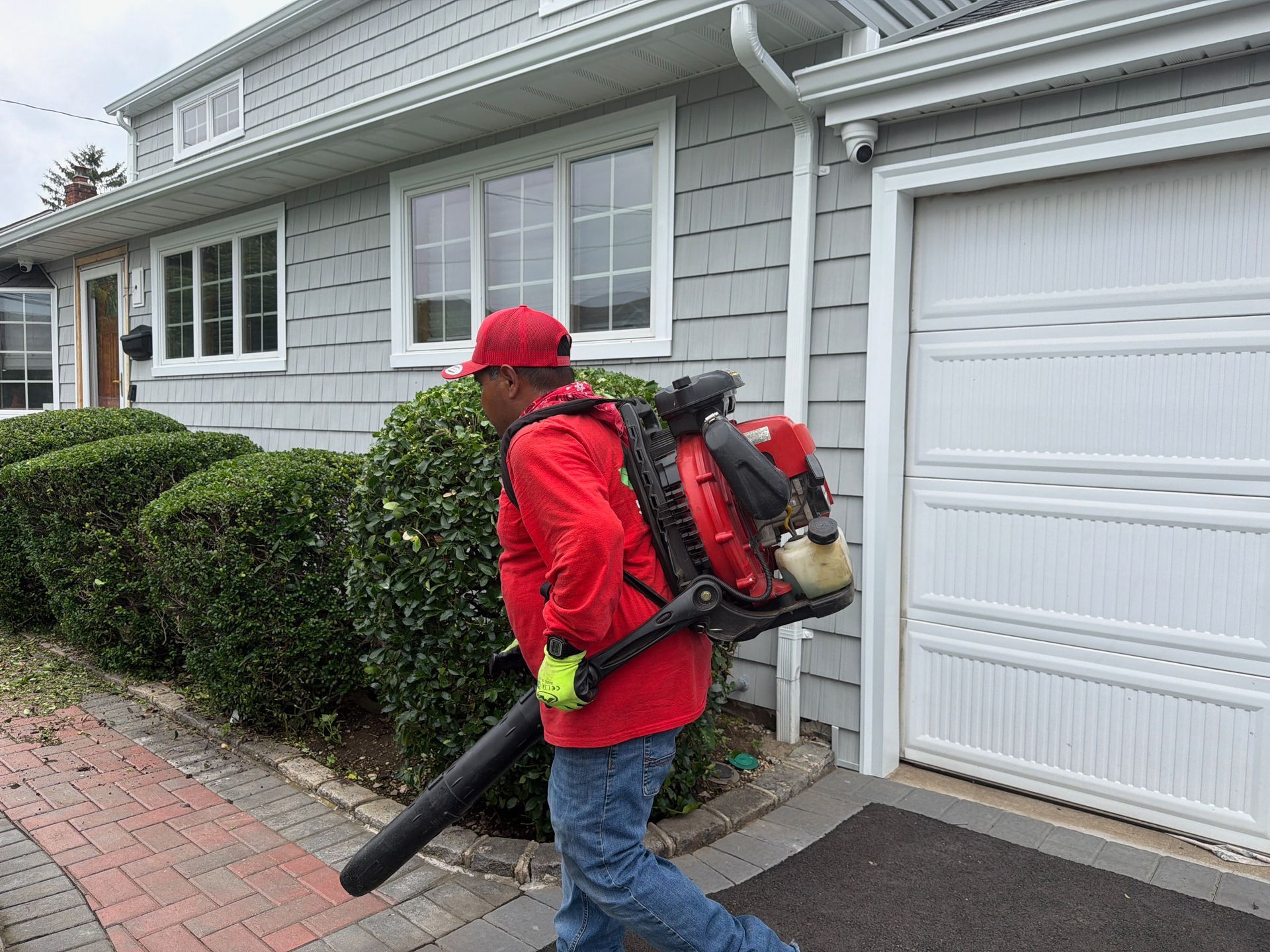 Worker using a backpack leaf blower on a sidewalk beside a house and garage.