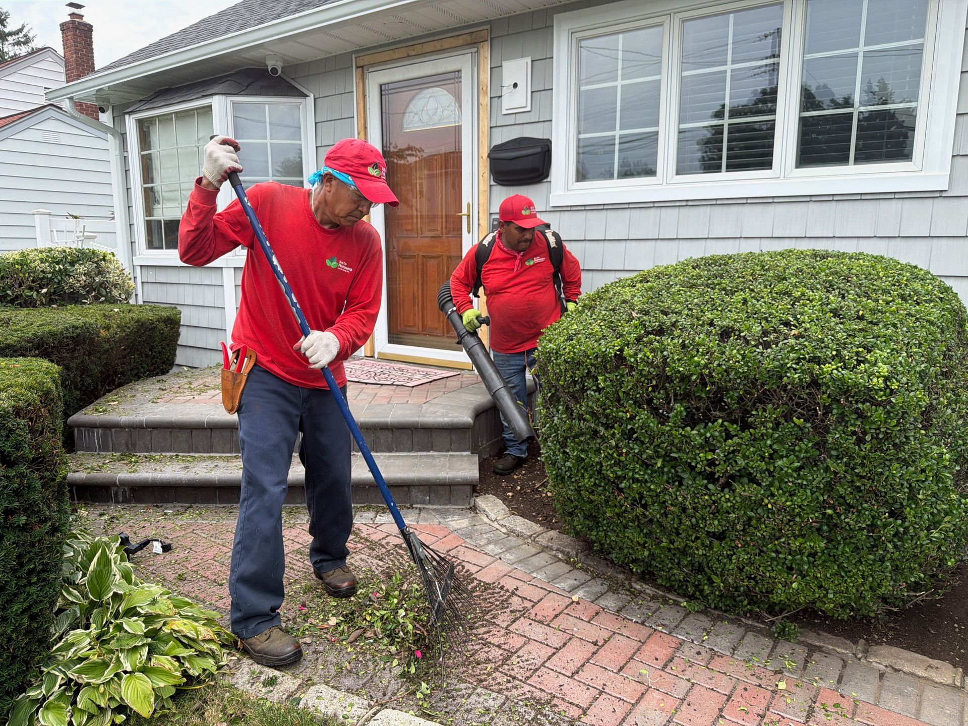 Two workers in red jackets and caps use leaf blowers on a brick walkway outside a house.
