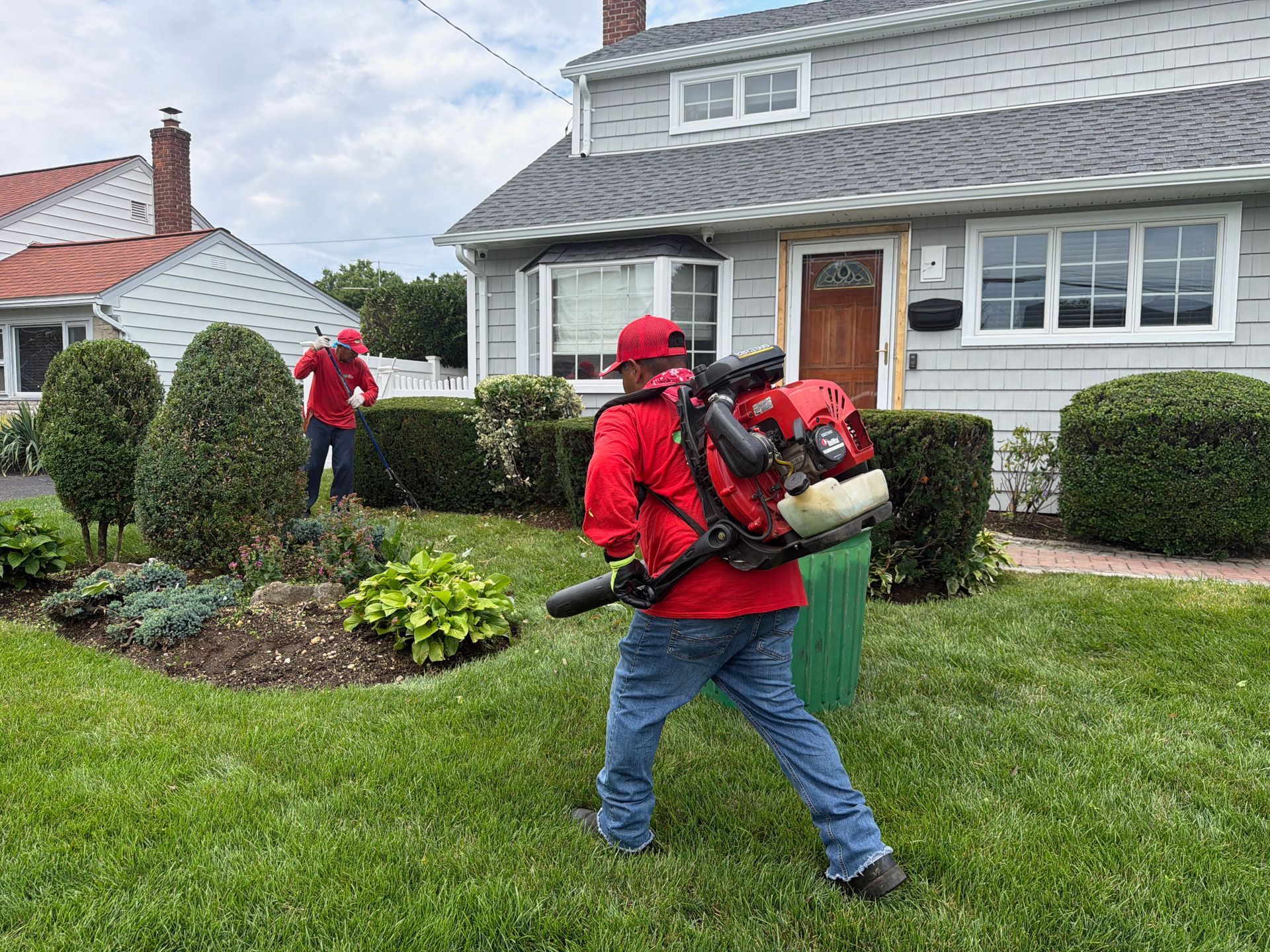Two workers in red gear using leaf blowers to clear a suburban front yard and garden bed