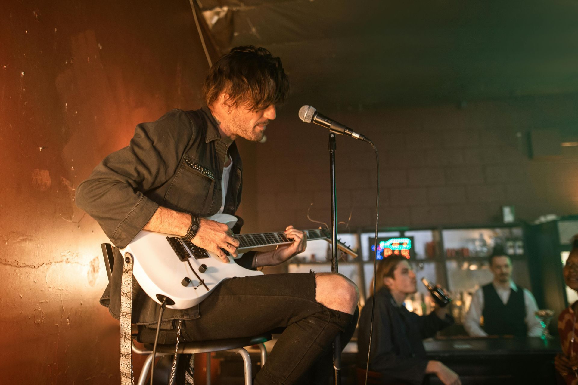 A man is sitting on a stool playing a guitar and singing into a microphone.