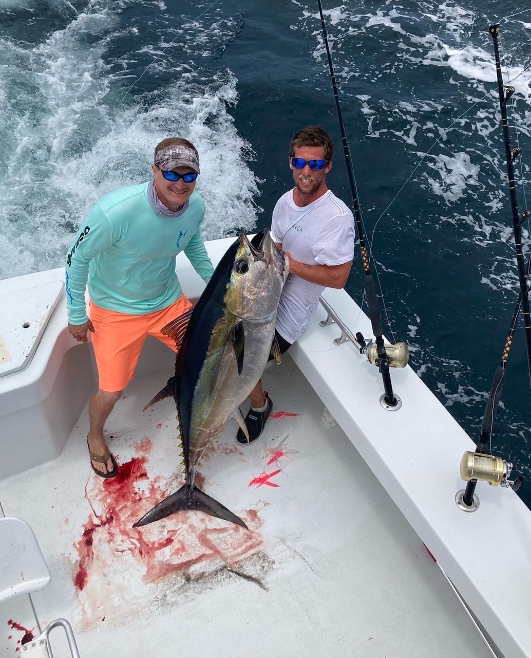 Two men on a boat hold a large tuna fish, blood on the deck. Ocean water in background.