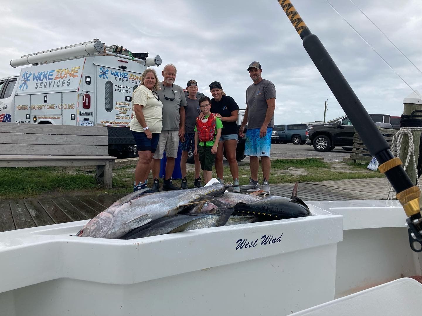 Group stands on a boat with caught fish. Cloudy sky, truck in background.