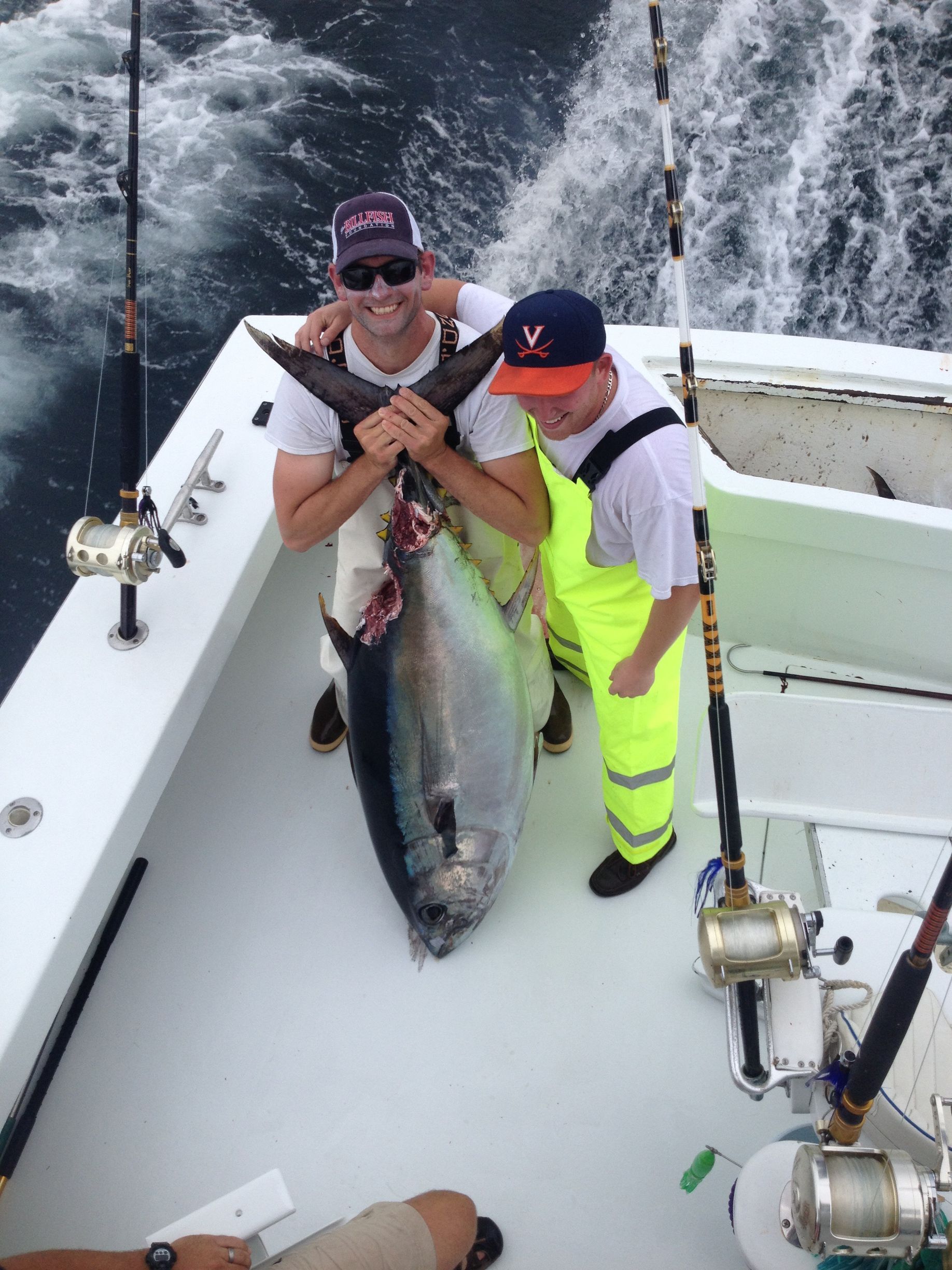 Two men on a boat, holding a large tuna they caught. Blue water, white boat, sunny day.