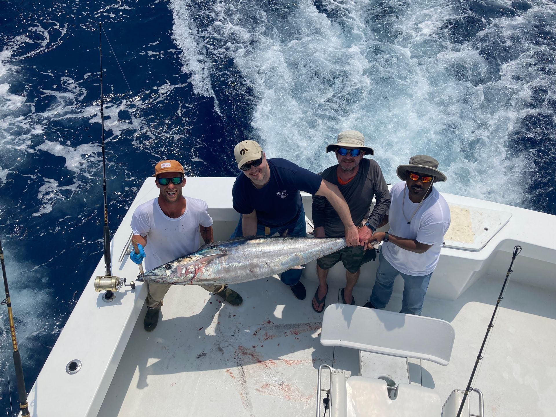 Four people on a boat holding a large fish they caught in the ocean; blue water and sky.