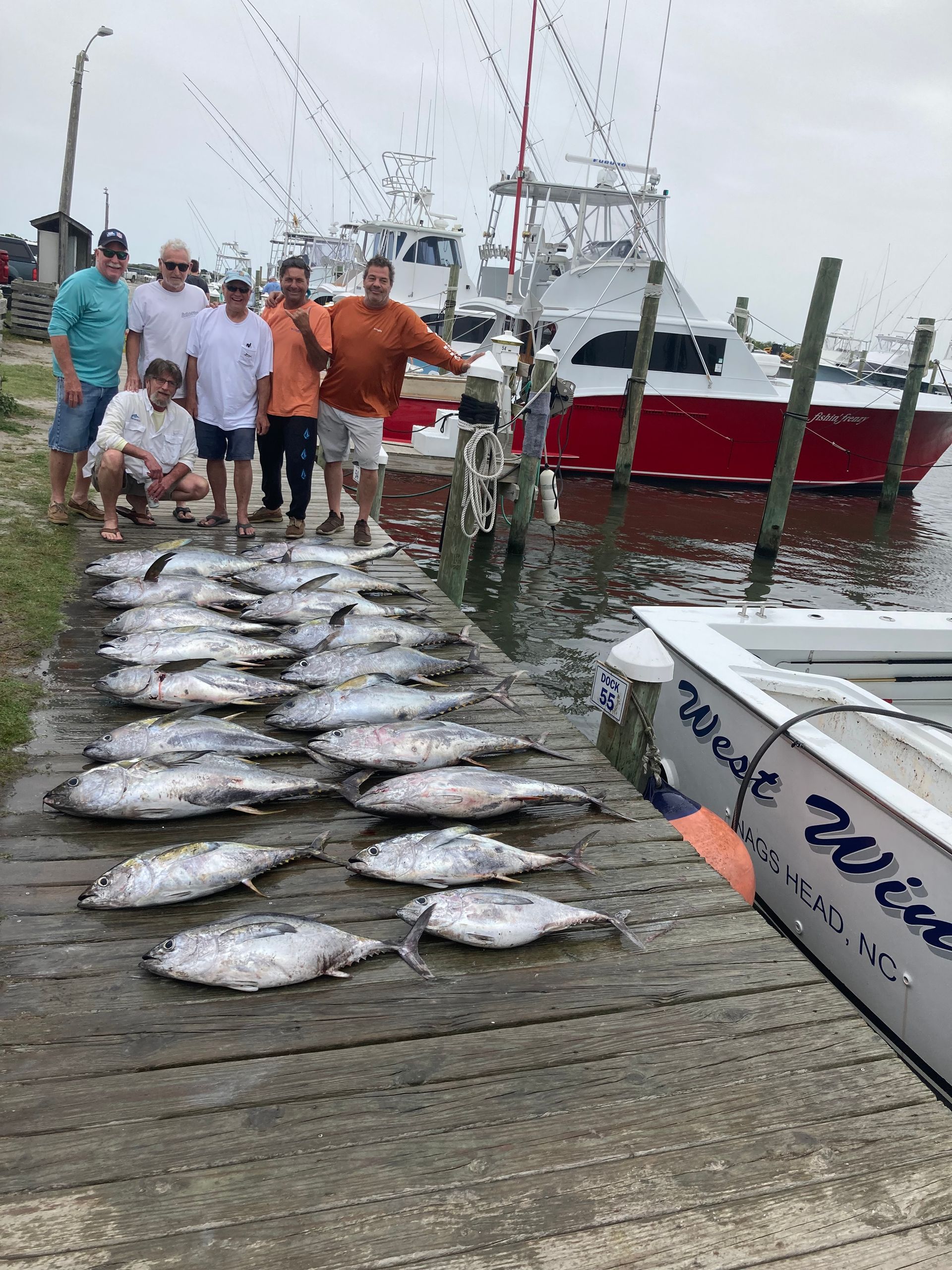 Group of people pose with a large catch of fish on a dock near boats.