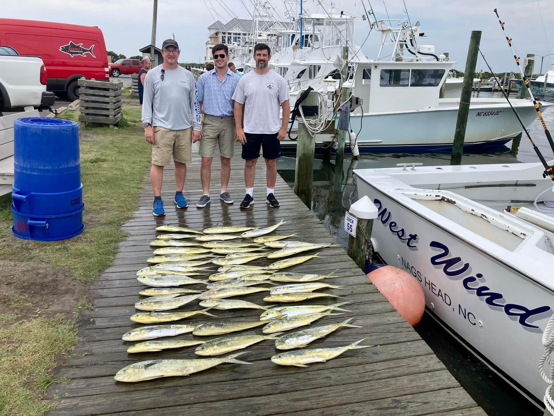 Three men stand on a dock with a large catch of fish; boats and ocean in the background.