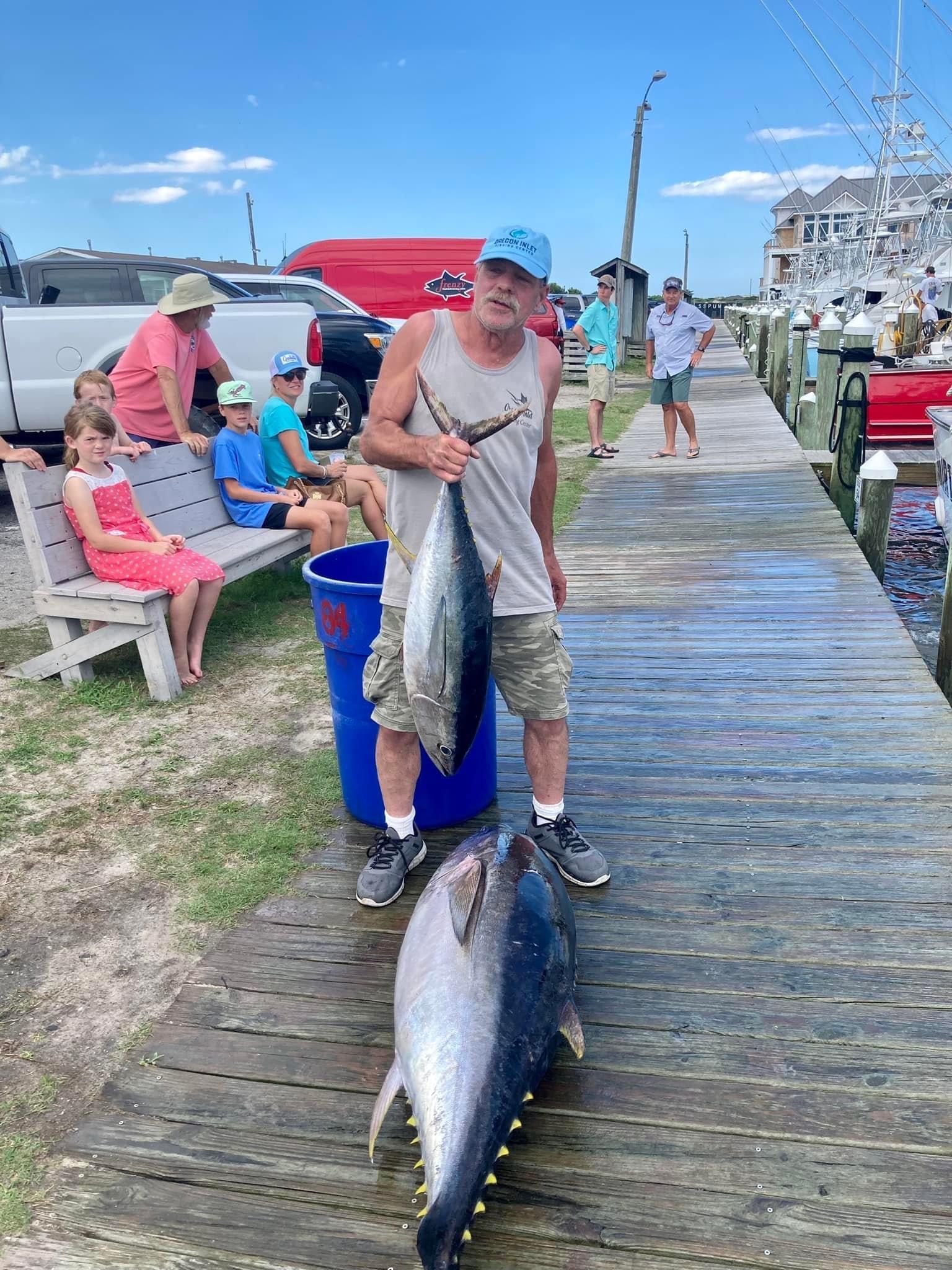 Man holds up fish on a pier; two other large fish lie below. People nearby, blue sky.