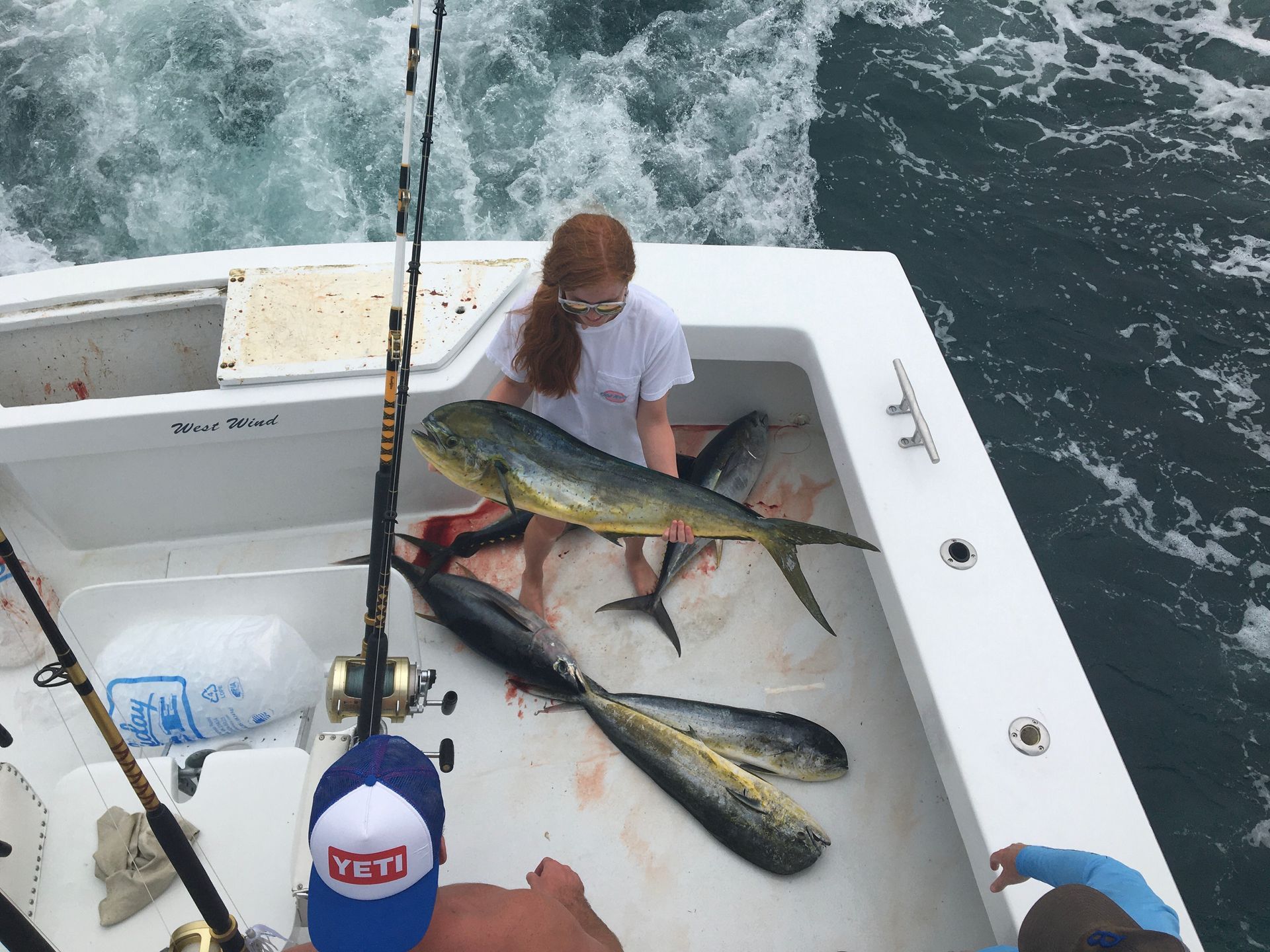 Woman holding a mahi-mahi on a boat, surrounded by other caught fish. Ocean background, fishing rods visible.