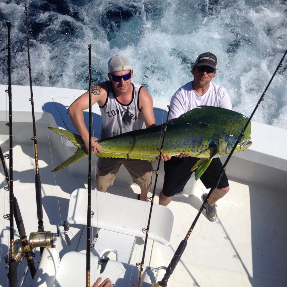 Two people on a boat holding a large, colorful fish, likely caught while fishing, with fishing rods visible.