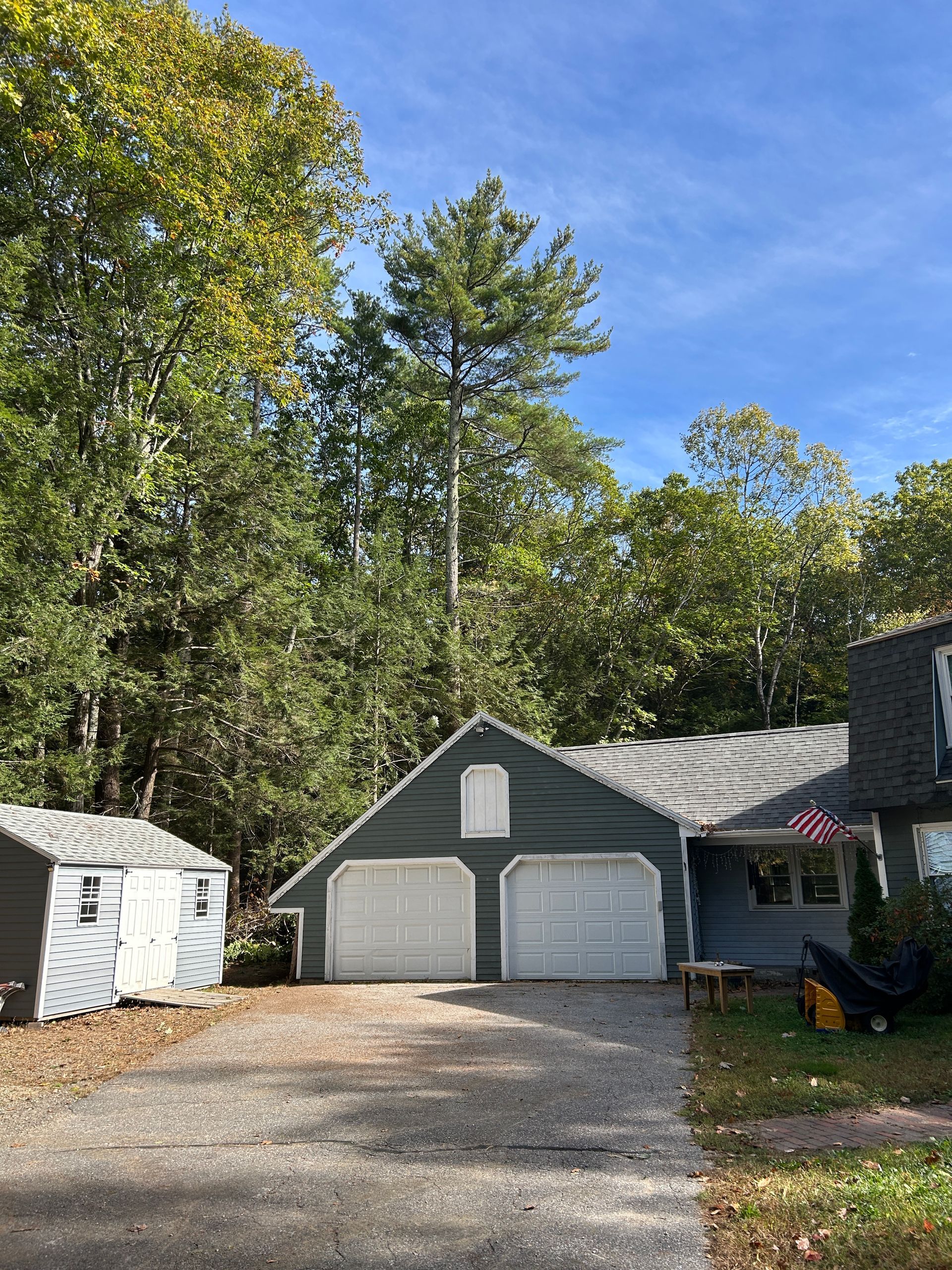 Two-car garage with white doors beside a small shed, gravel driveway, and trees under a blue sky.