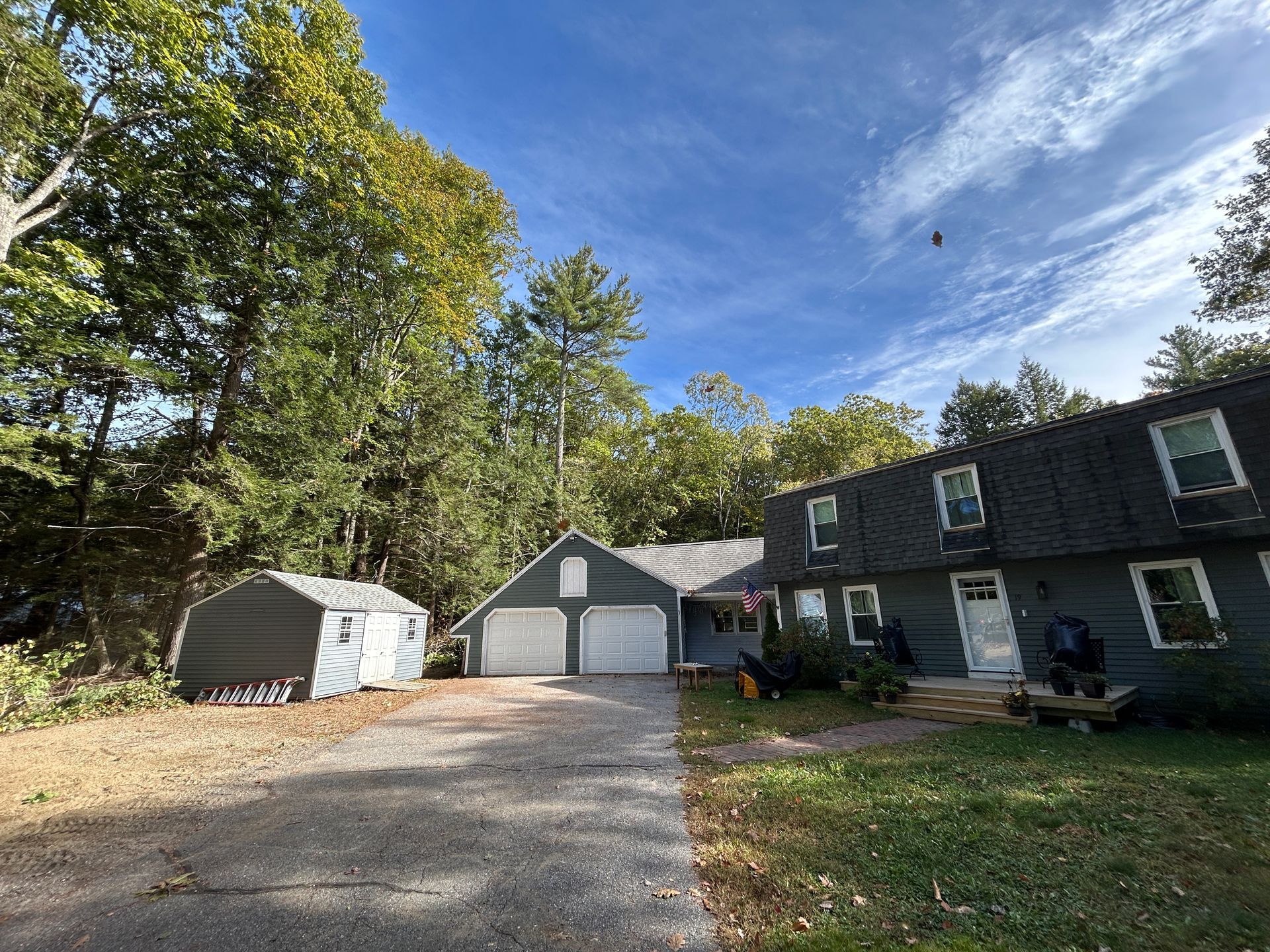 Suburban driveway with a garage, dark house, white shed, and trees under a blue sky