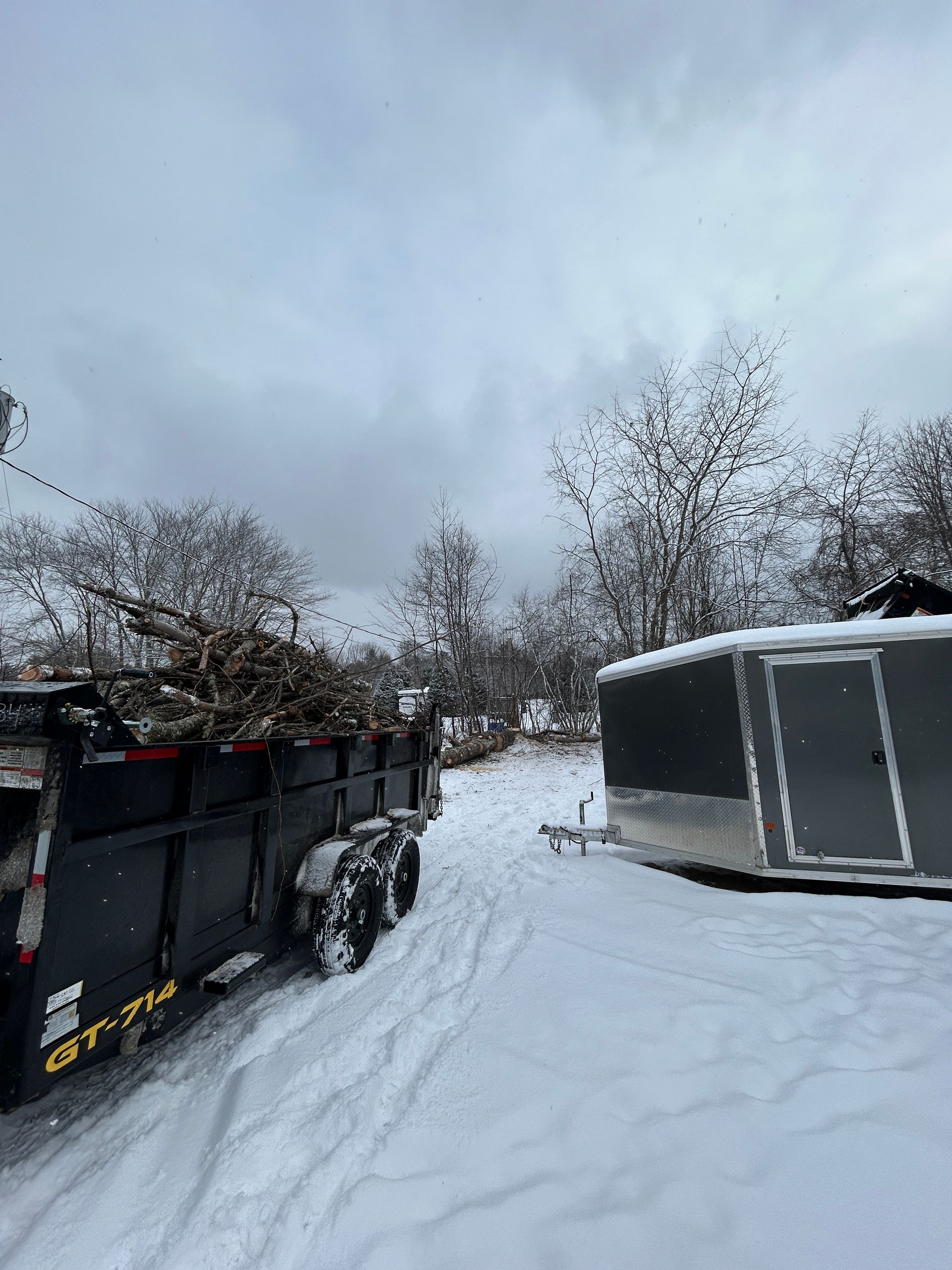 Black truck parked in snowy yard beside a gray trailer under an overcast sky