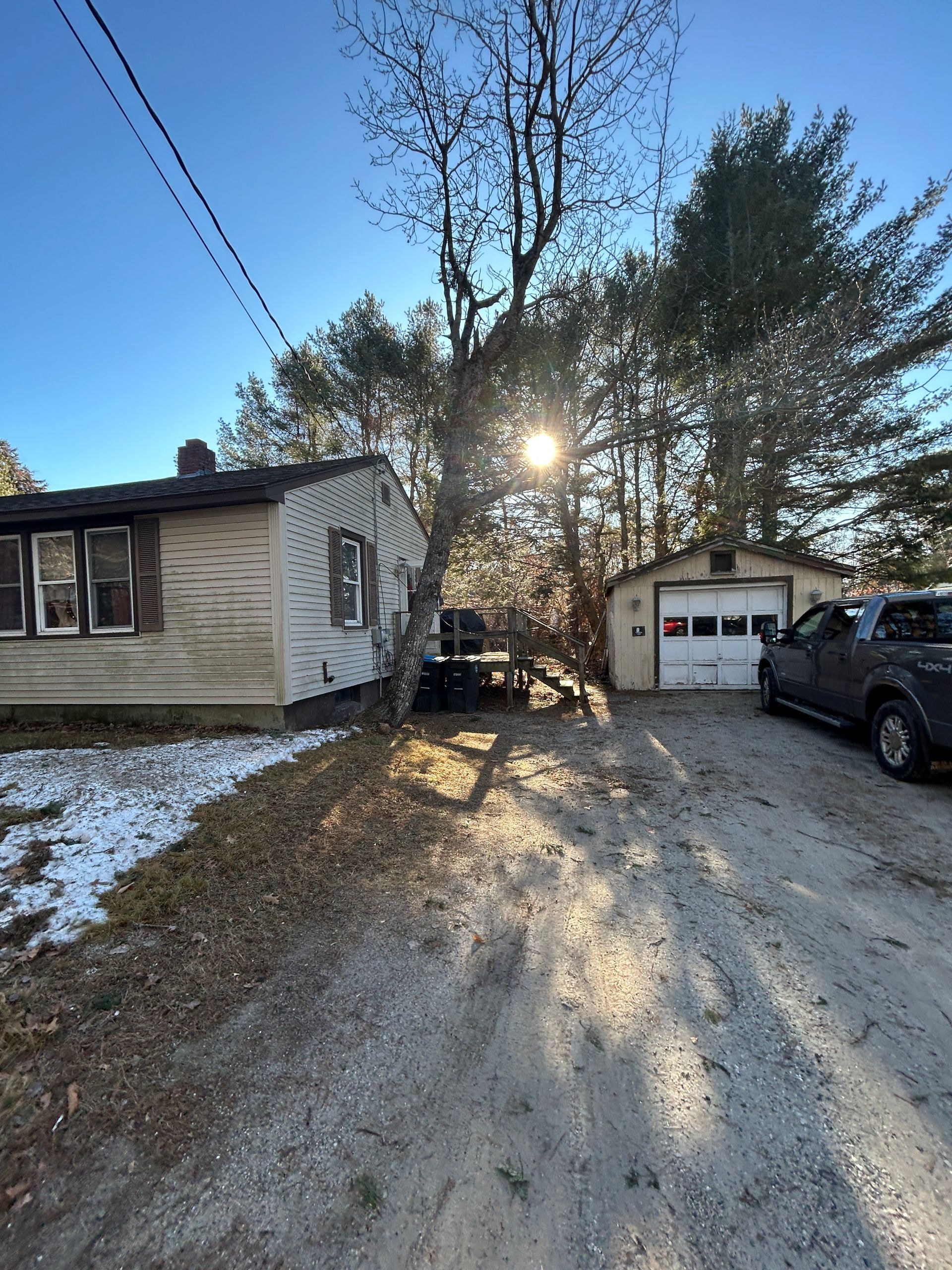 Snowy driveway between a small house and garage at sunset, with a parked SUV and bare trees.