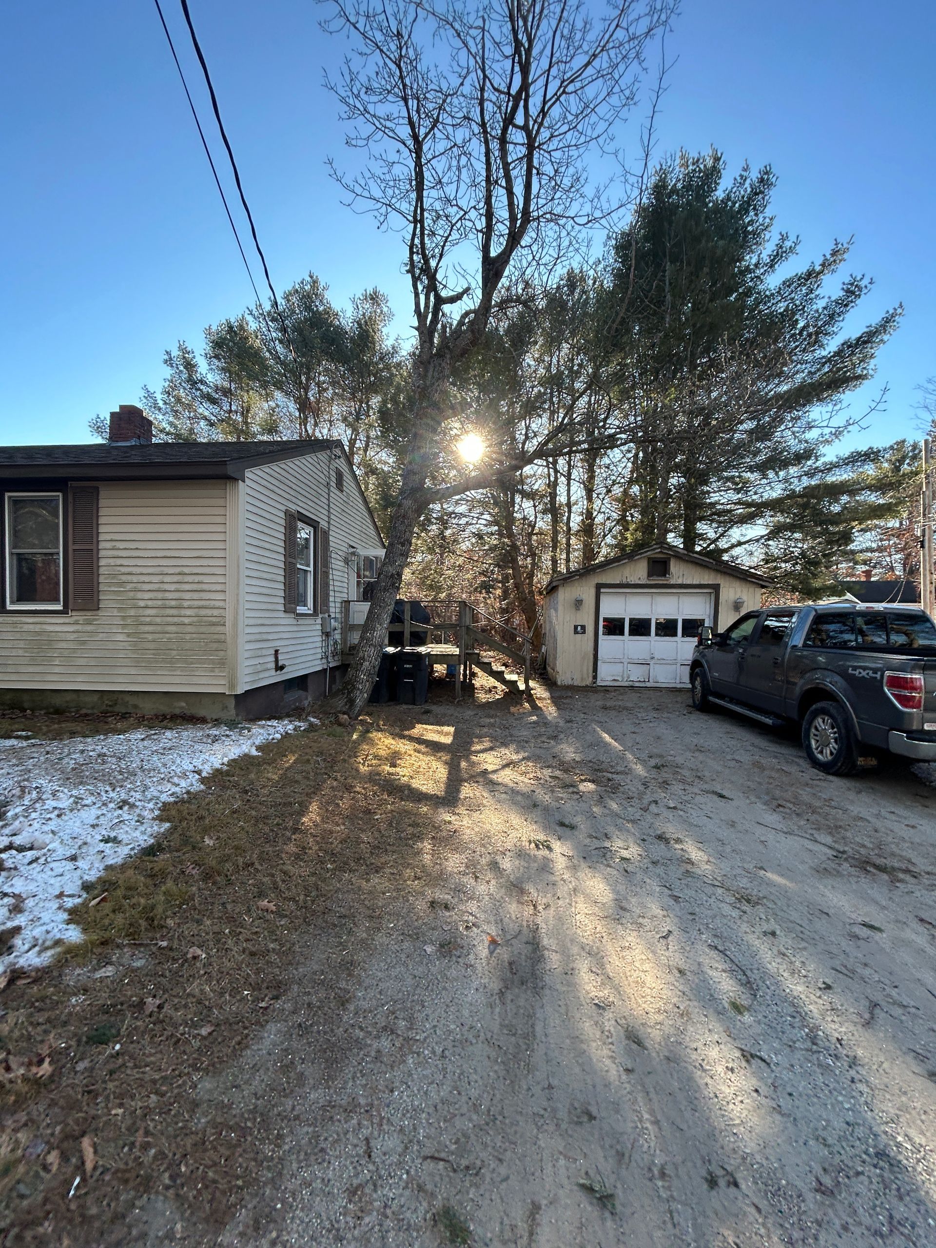 Snowy driveway beside a house and garage, with cars parked and sun shining through trees.