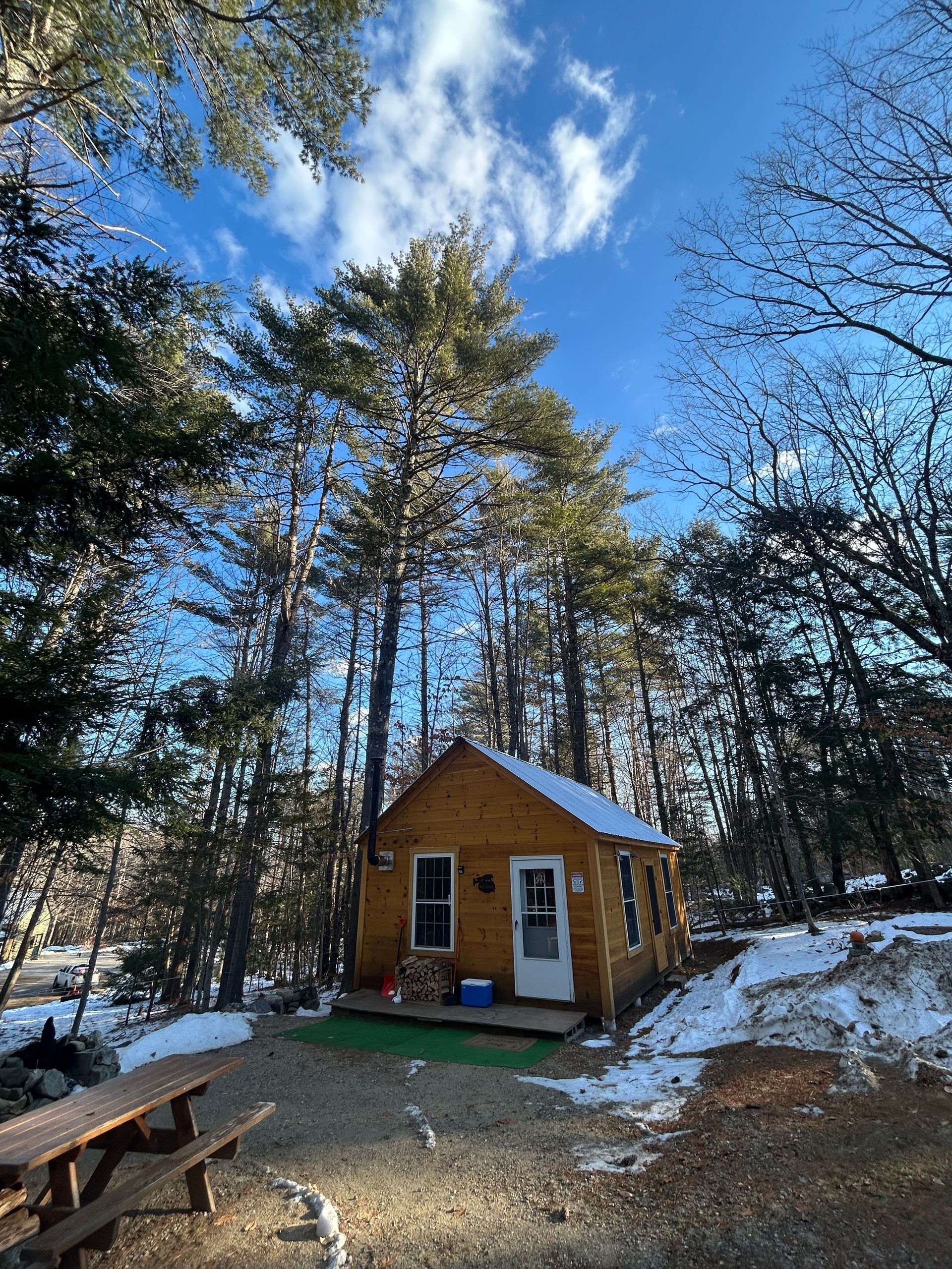 Small yellow cabin in a snowy wooded clearing under a blue sky, with picnic tables in the foreground.