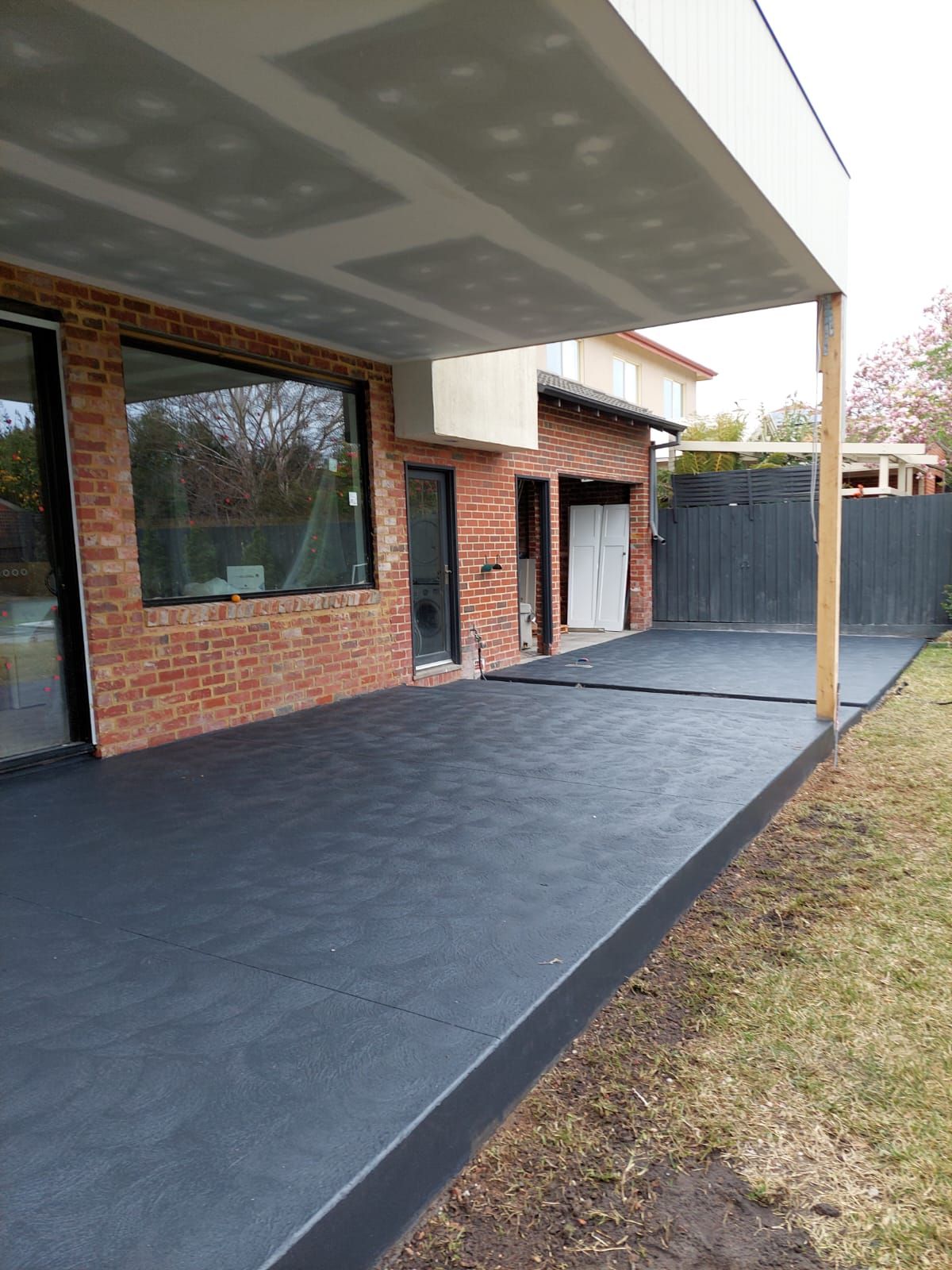 Covered patio with brick house walls, dark flooring, and a fenced yard outside.