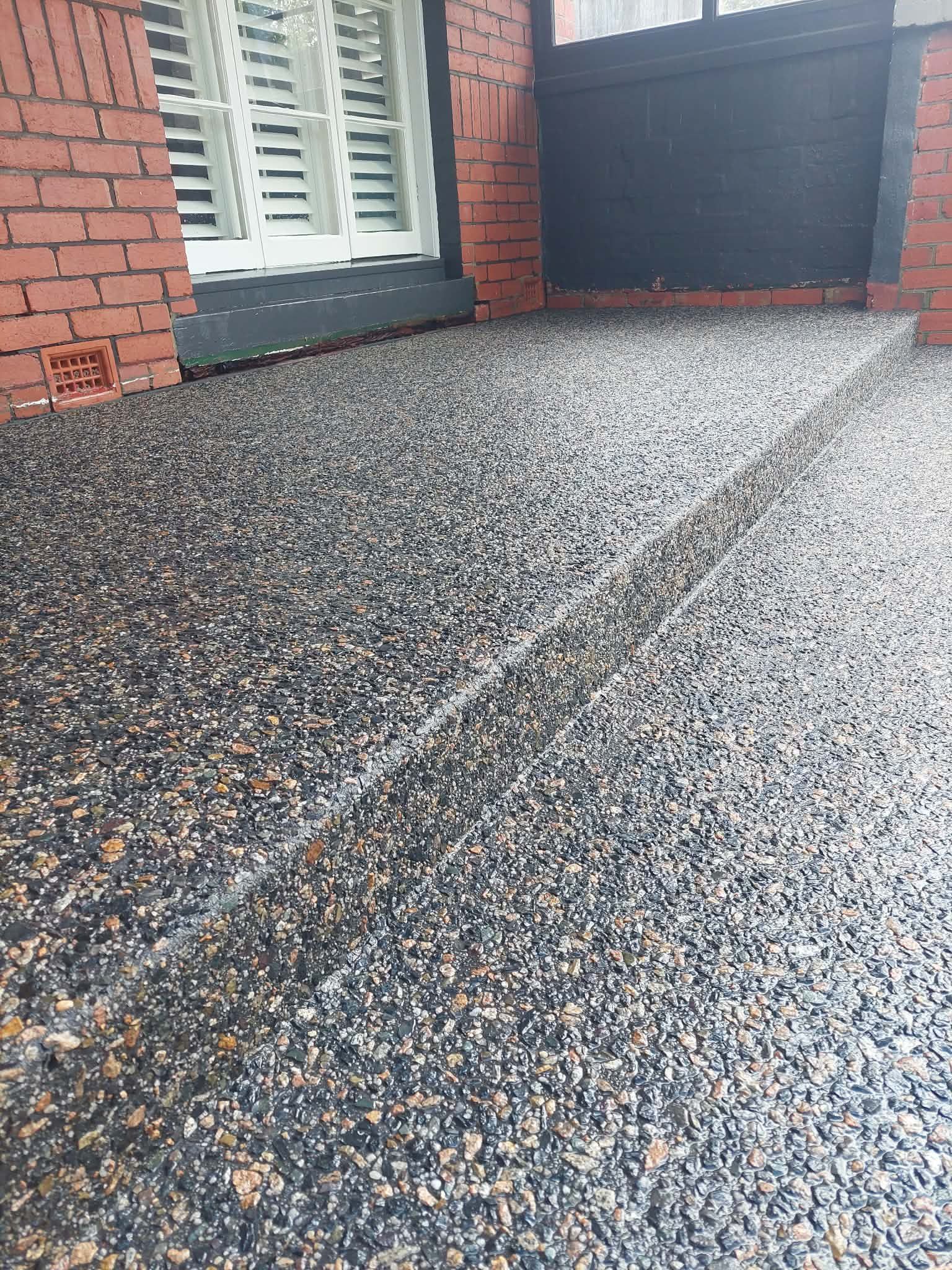 Gravel-textured driveway beside a red brick house and dark garage door, with white-trimmed windows nearby