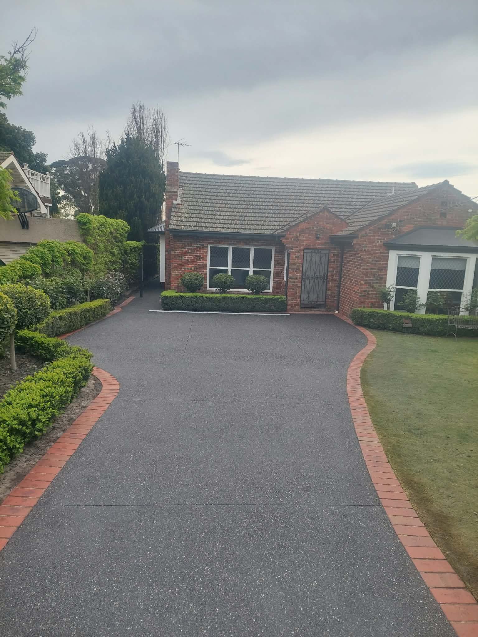 Brick house with a curved dark driveway and brick edging, framed by green hedges and an overcast sky.