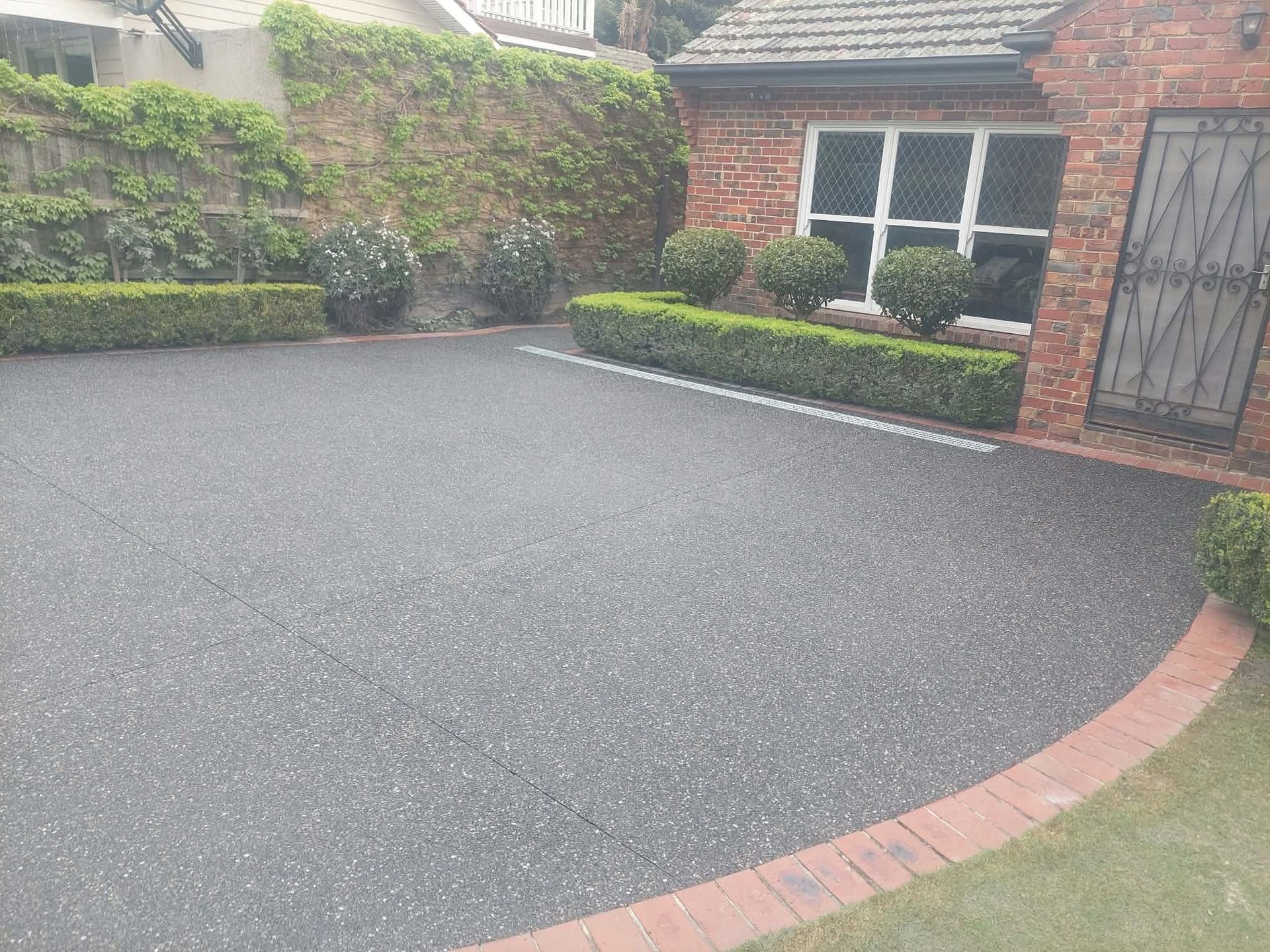Freshly paved dark driveway beside a brick house, with hedges, windows, and a curved red brick border.