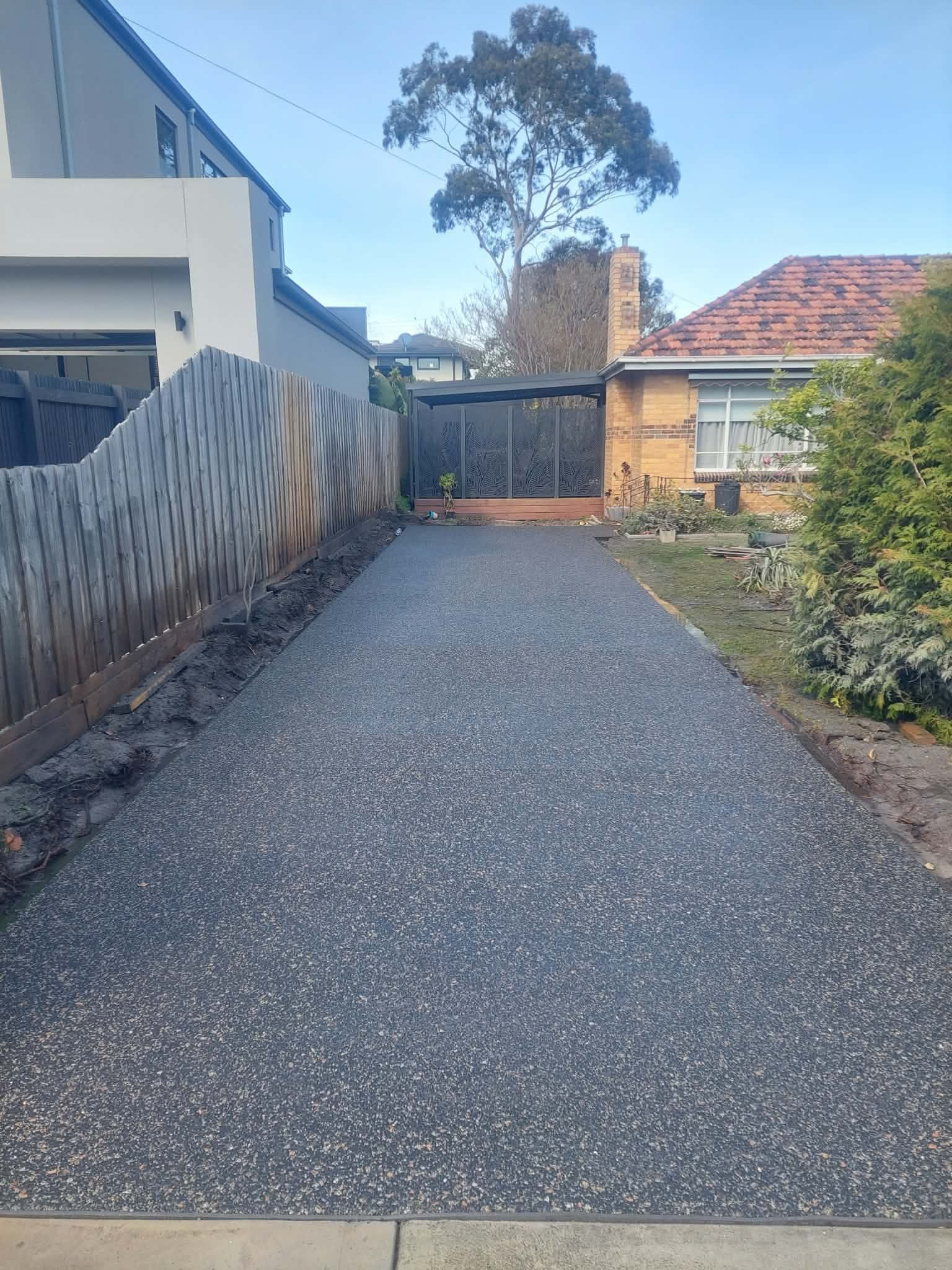 Gravel driveway between a modern house and fence, leading to a gate and brick house in the distance.
