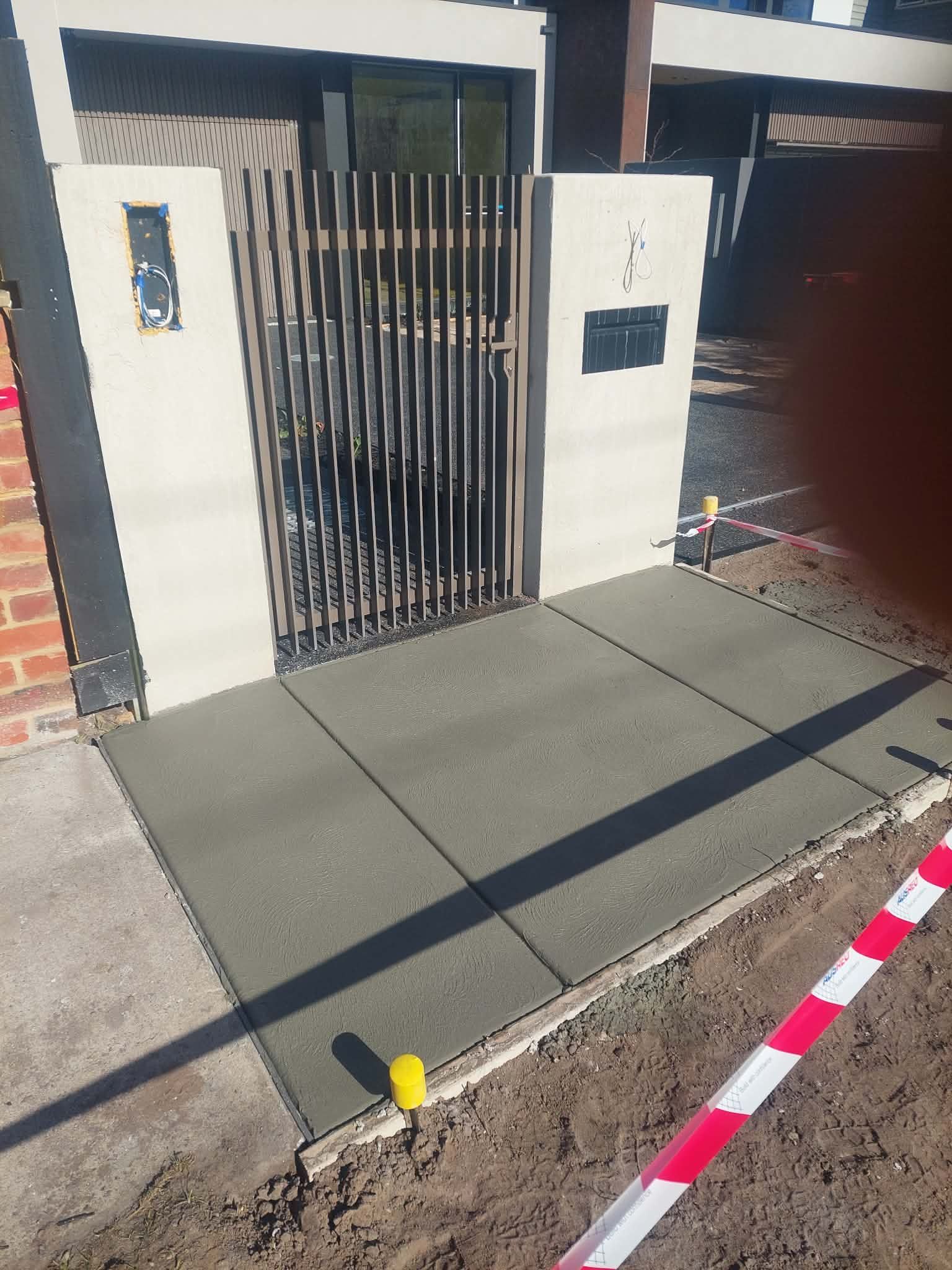 Front gate with black metal bars and a concrete sidewalk ramp beside a brick house.