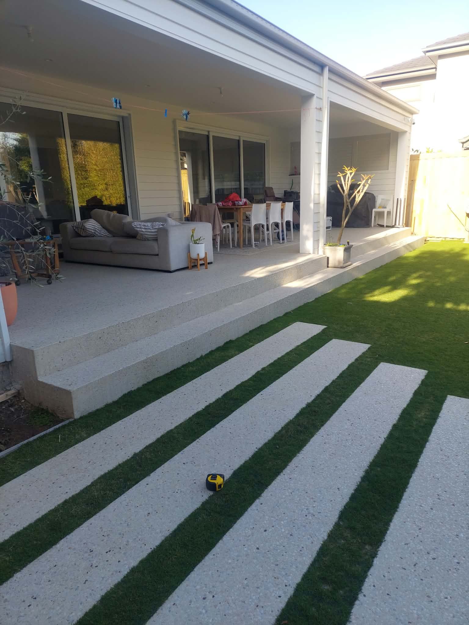 Backyard patio with sofa, dining set, and striped lawn path leading to a modern house.