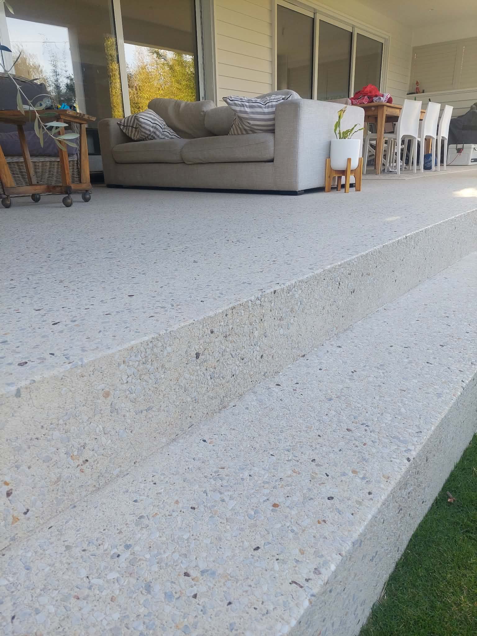 Front porch with wide concrete steps, gray sofa, white chairs, and a glass door beside a patch of grass