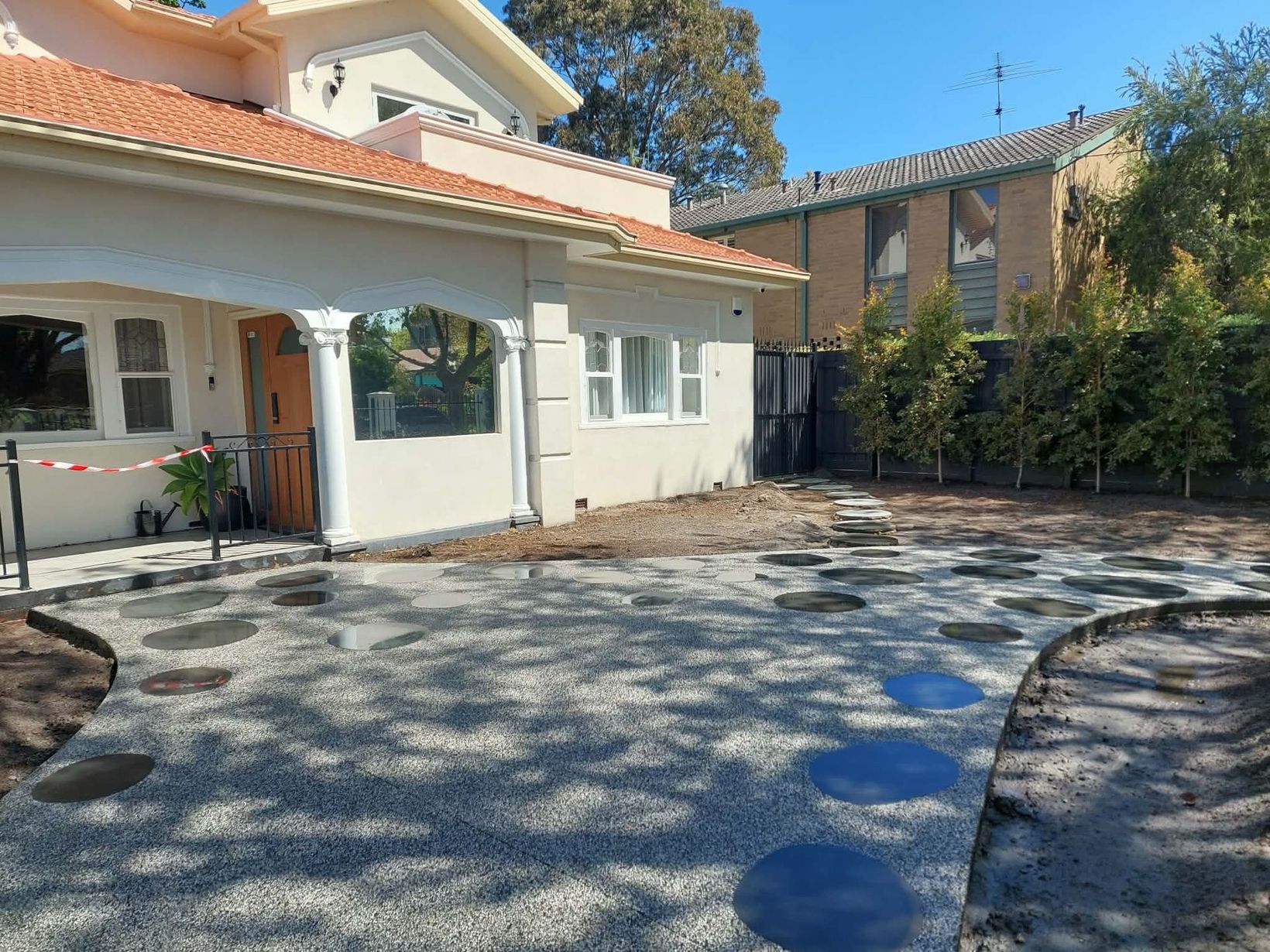 Sunlit driveway beside a house with a tiled roof, white walls, and trees casting shadows