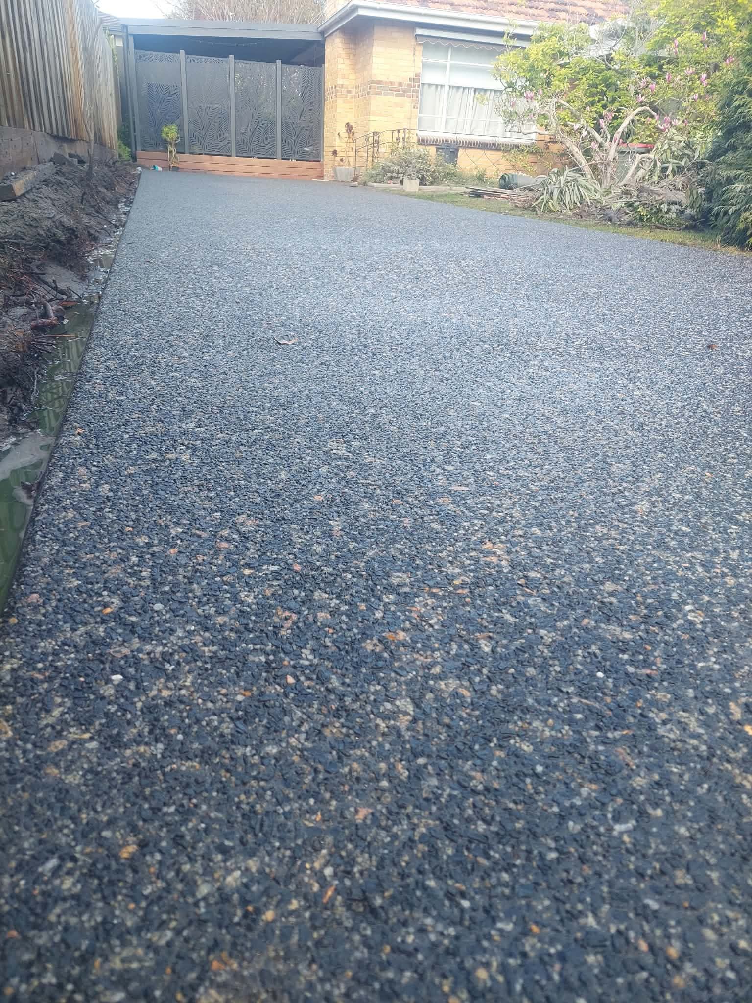 Gravel driveway leading to a wooden gate, with shrubs and a house in the background.