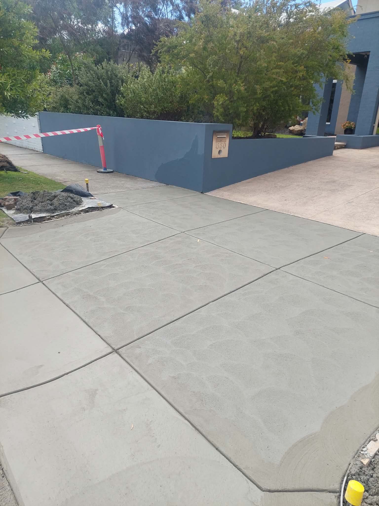Concrete sidewalk beside a blue-gray retaining wall, with a taped-off area and trees in the background.