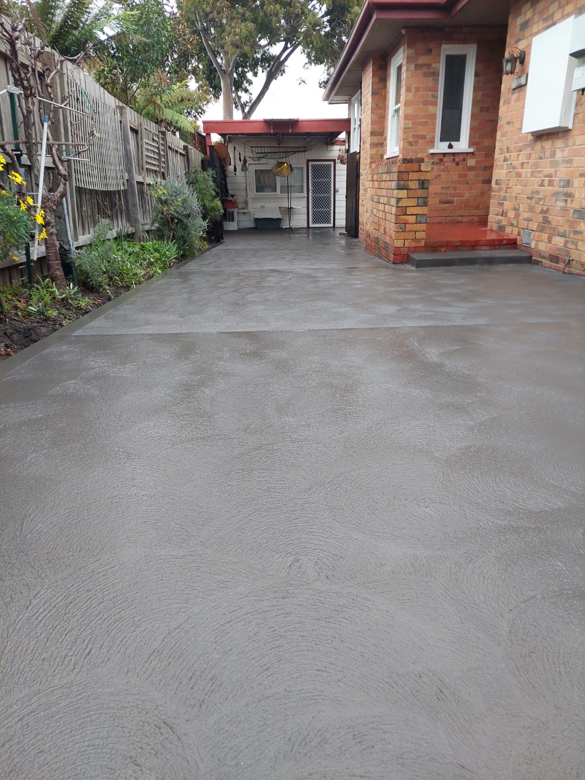 Concrete driveway beside a brick house, leading to a covered entry and garden fence.
