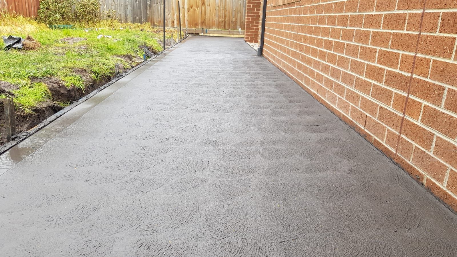 A freshly paved driveway between houses, with wooden fences and trees lining the sides.