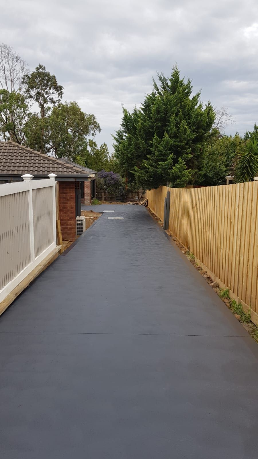 Wet residential driveway between white and wooden fences, leading to trees under an overcast sky