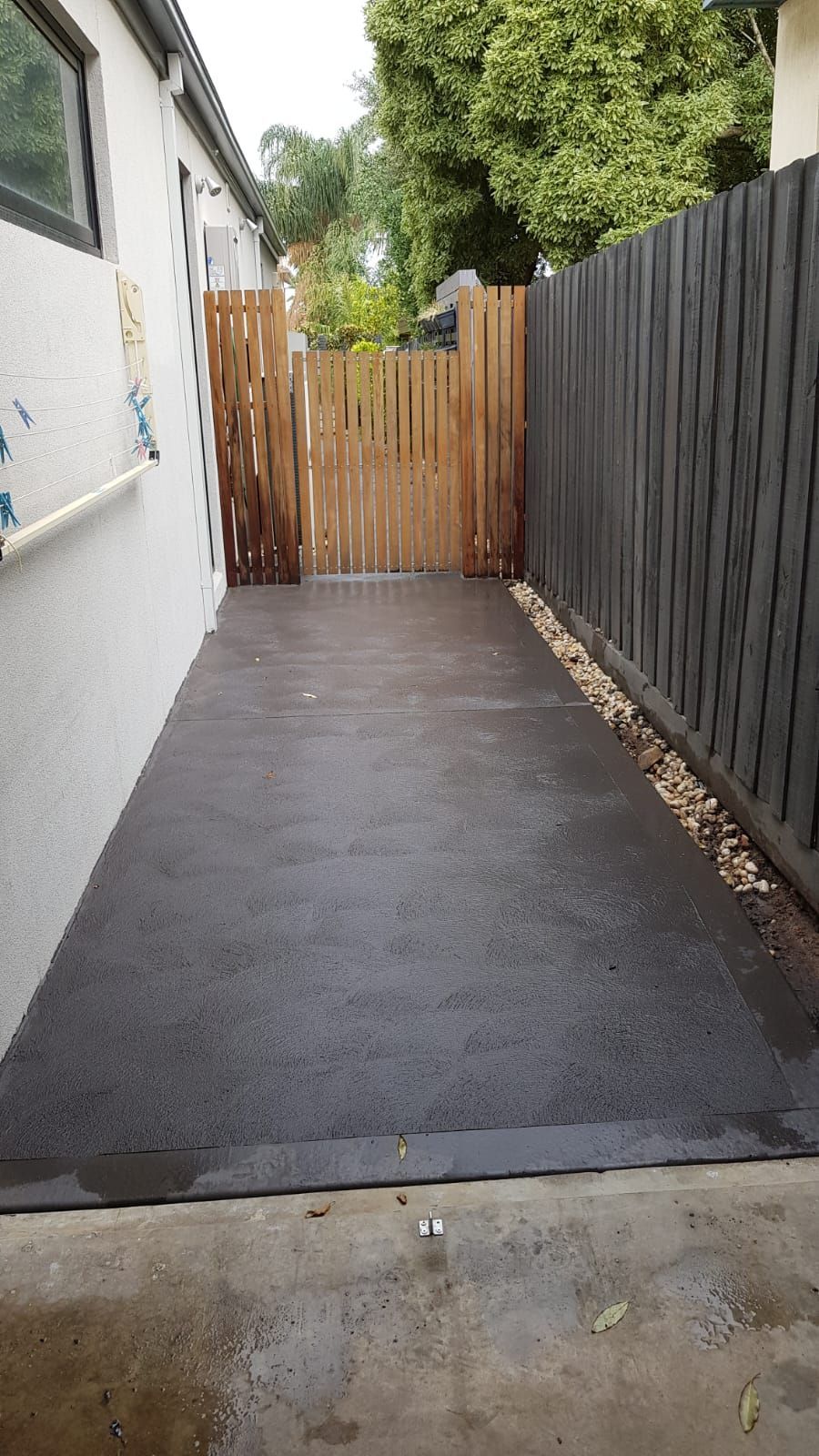 Narrow paved side yard with a white wall, black fence, and wooden gate at the end.